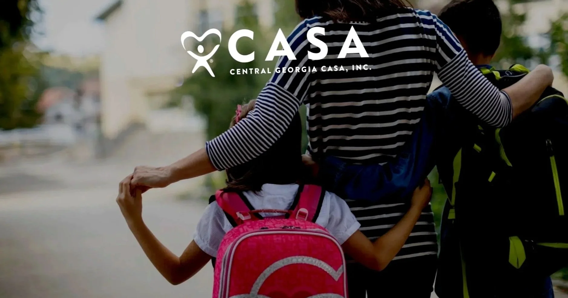 Two children, a boy and a girl, walking outdoors with backpacks, holding hands, and the girl has her arm around the boy's shoulders. The scene appears to be in a neighborhood or park setting.
