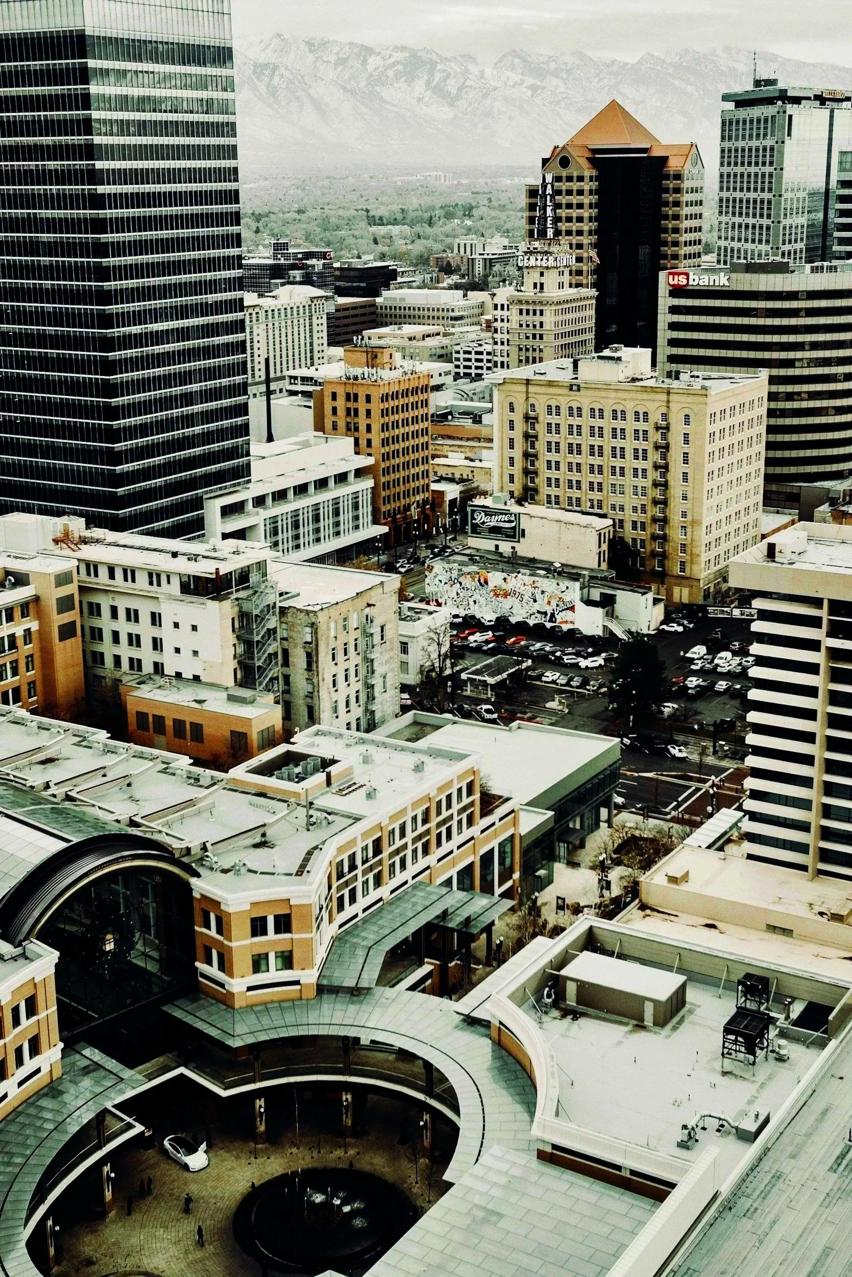 A cityscape view showing tall skyscrapers, buildings, and streets with parked cars, mountains in the background, and a cloudy sky.