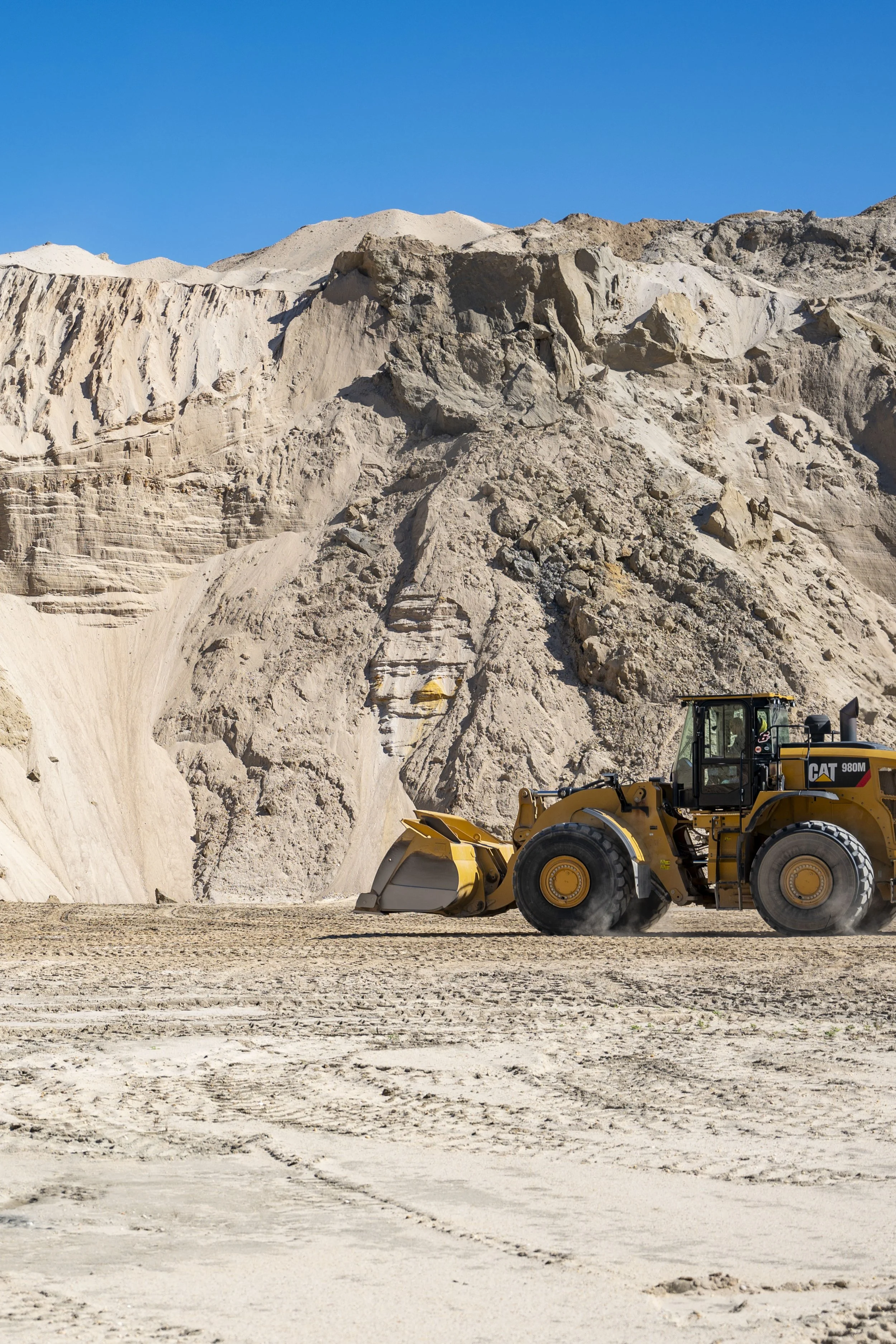 Yellow bulldozer working on a sandy construction site with large light-colored rocky hills in the background under a clear blue sky.