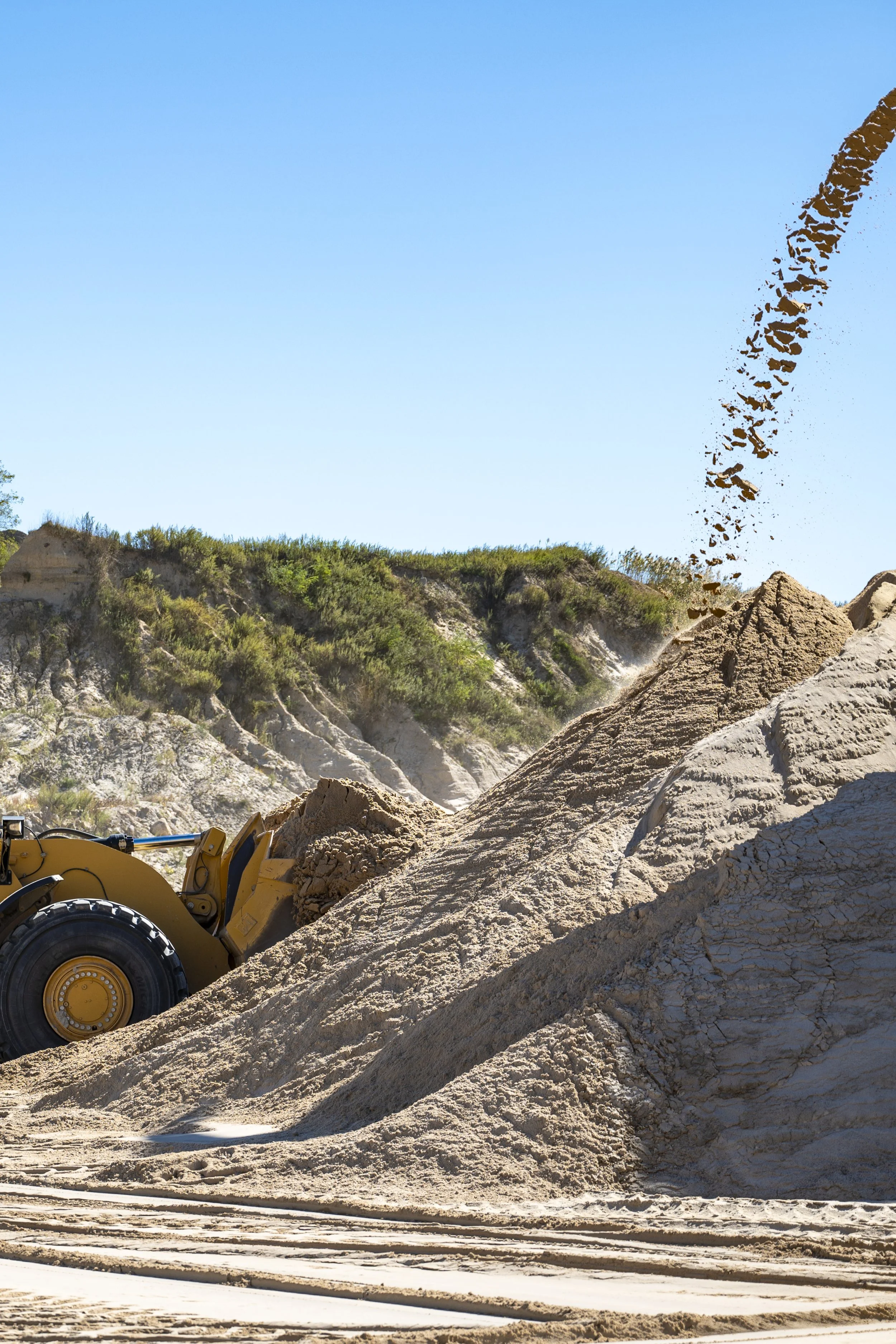A front loader bulldozer moving sand or dirt at a construction site with a hillside and blue sky in the background.