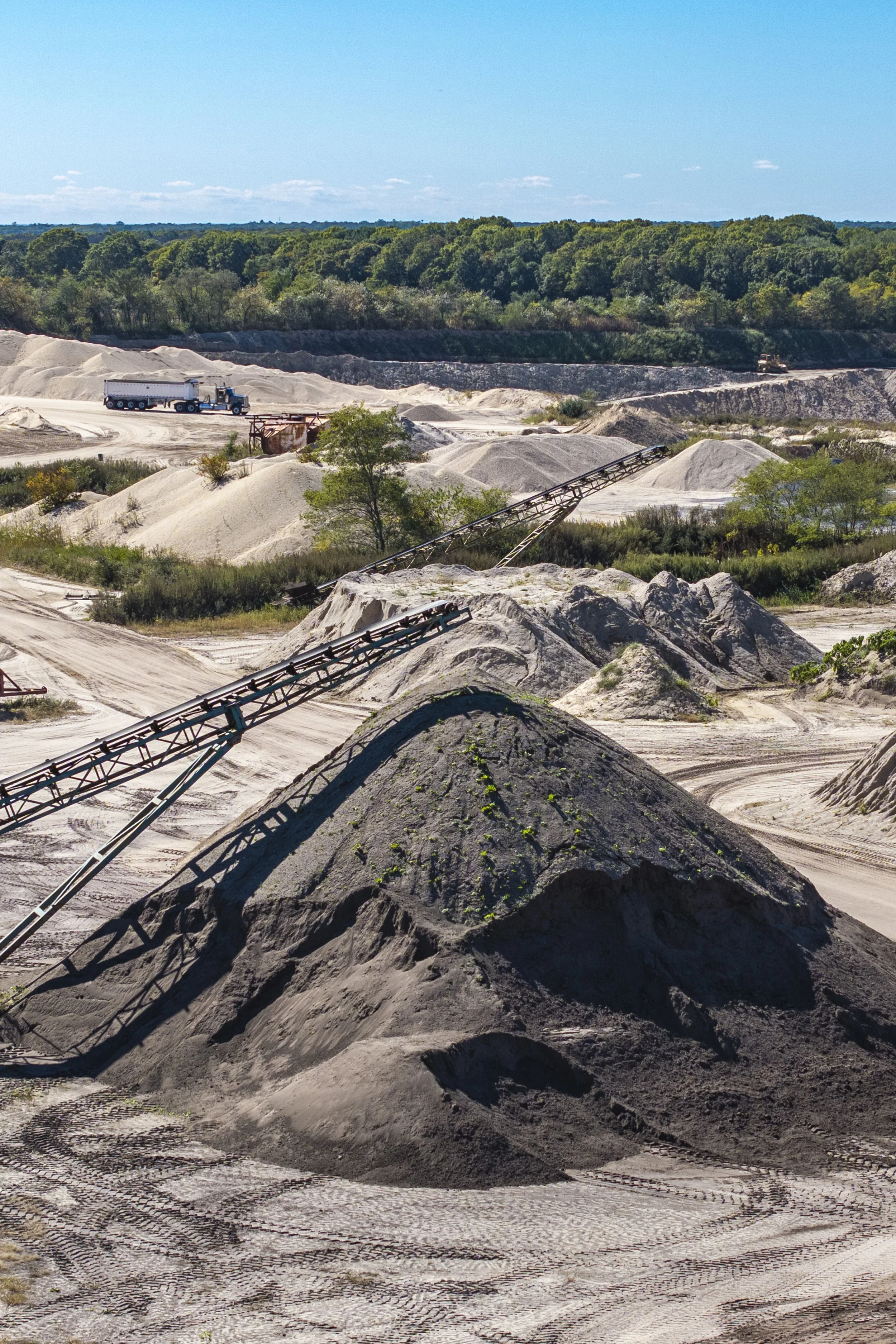 A large industrial sand or gravel mining site with piles of extracted materials, conveyor belts, and trucks under a blue sky.