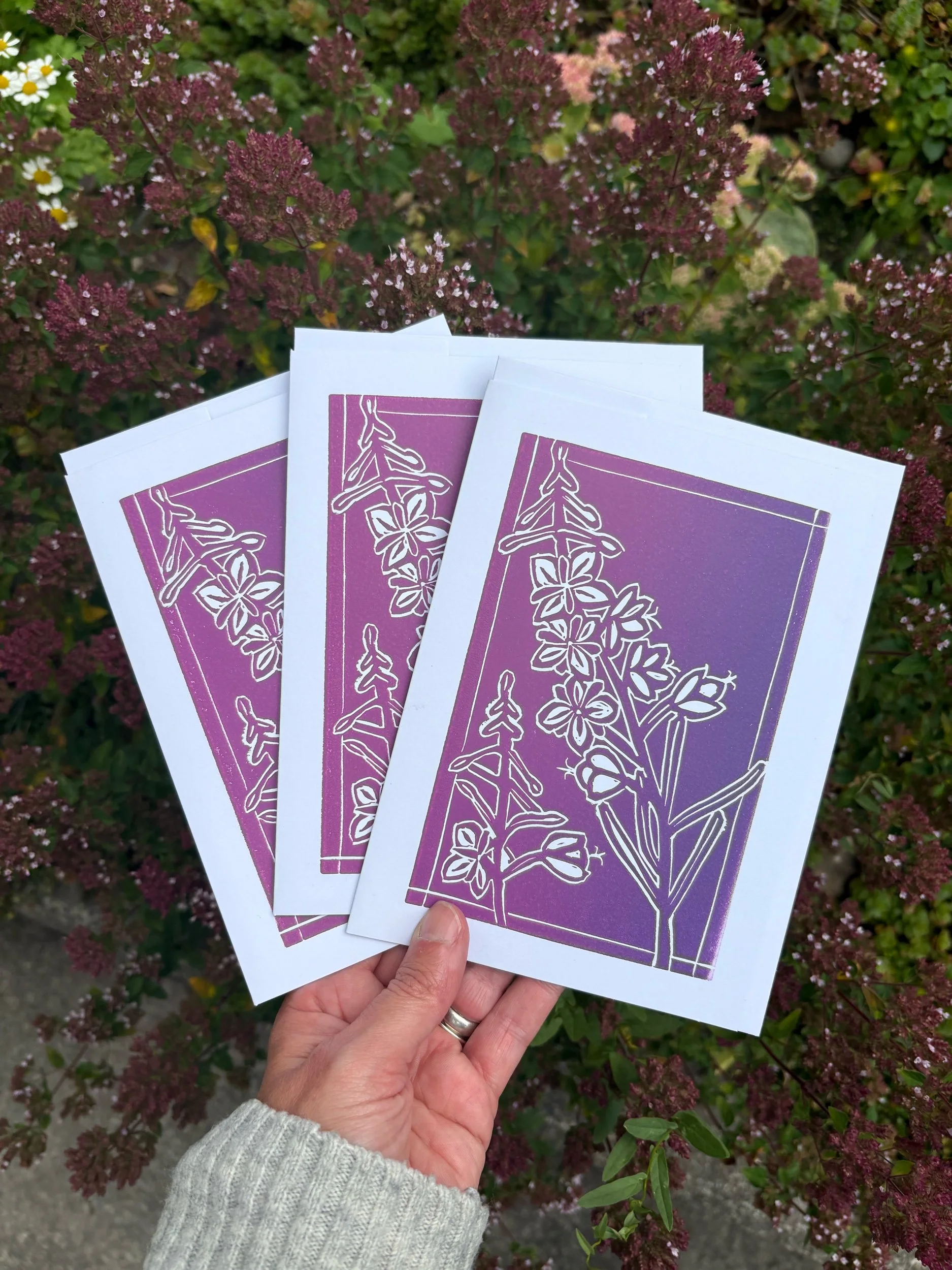 Hand holding three postcards featuring a stylized portrait of a woman with a halo and striped hair, set against foliage with red and green leaves.