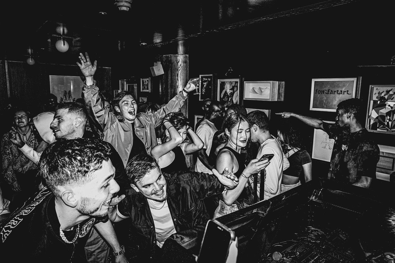 Crowd of young people dancing and enjoying music at a party or club, with some looking at their phones and others talking or dancing, in a dark room with framed artwork on the walls.