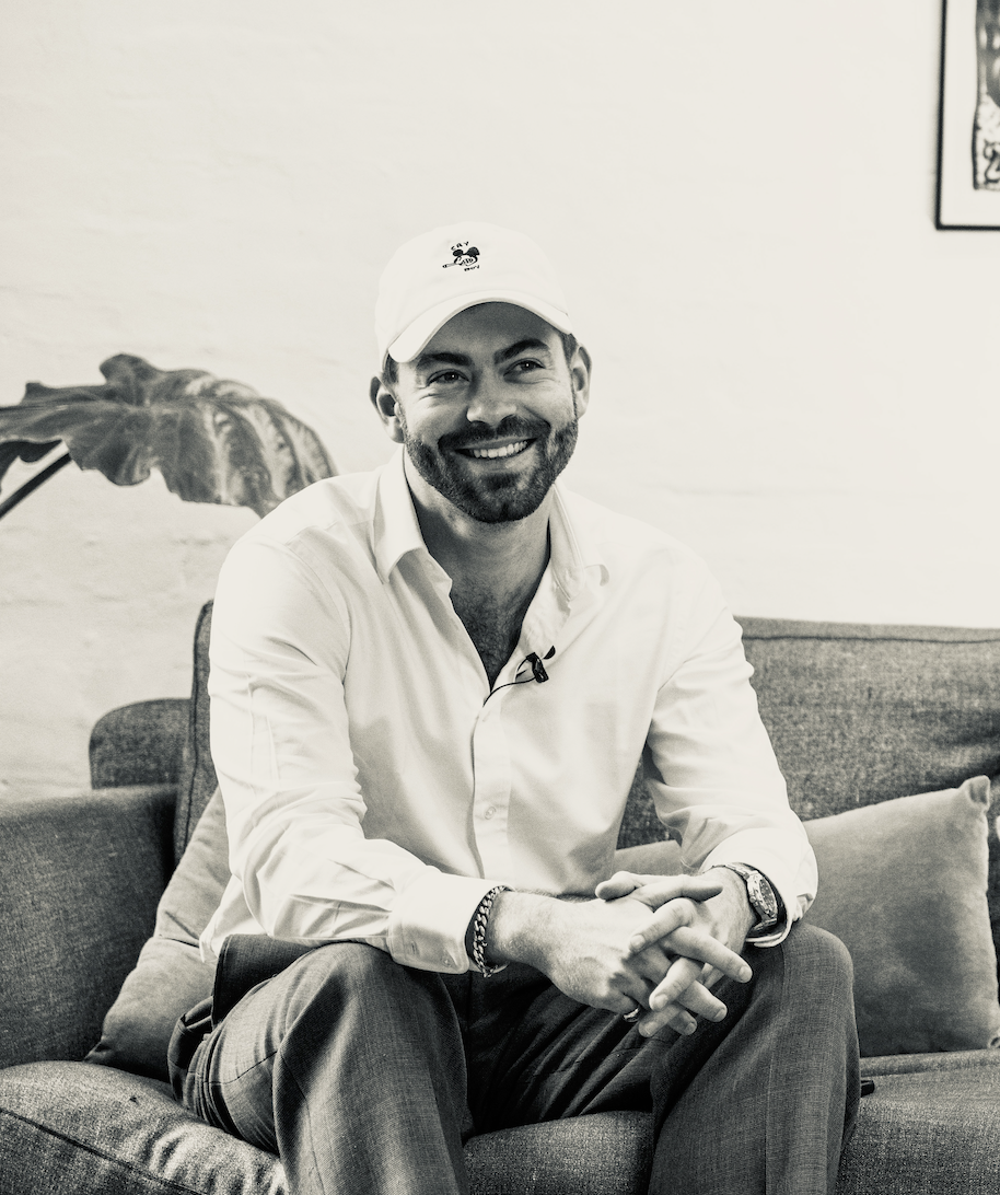 A man sitting on a sofa, smiling, wearing a white shirt, a white cap, and accessories, in a room with artwork and plants.
