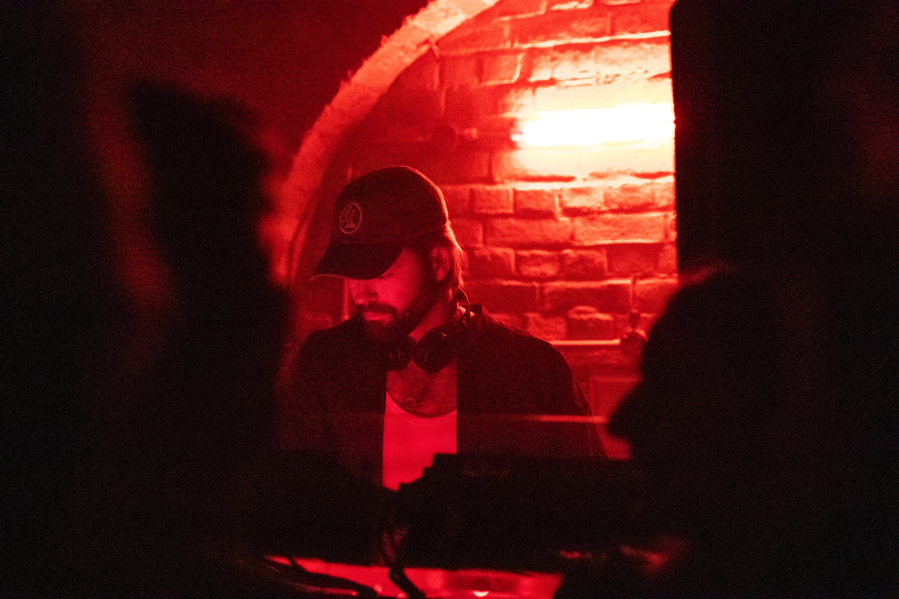 A DJ with headphones around his neck and a cap, performing in a dimly lit venue with red lighting and a brick wall background.