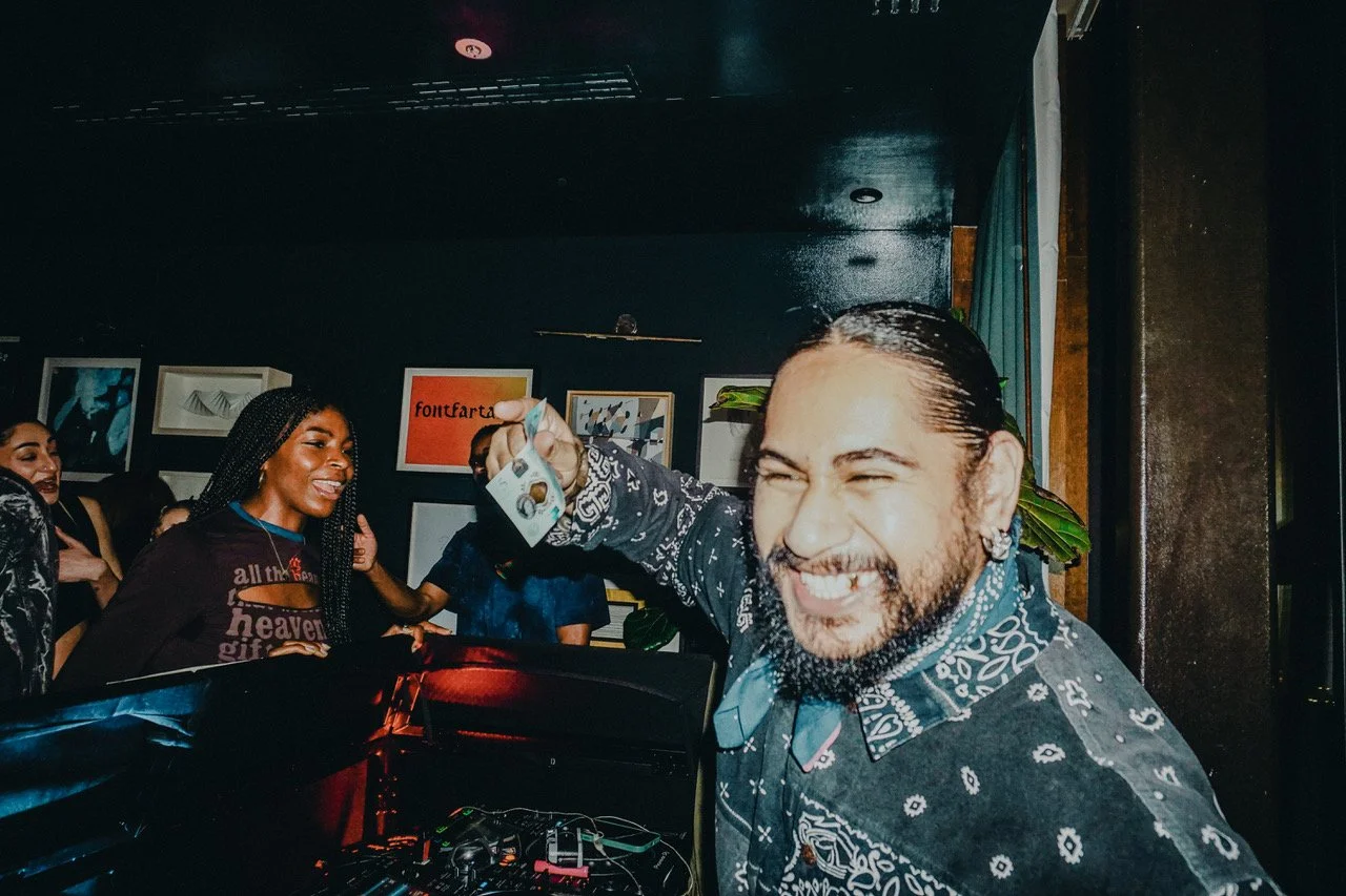 A man with a dark patterned shirt and long hair tied back smiling and holding money, with a group of women in the background laughing in a dimly lit room decorated with framed artwork.