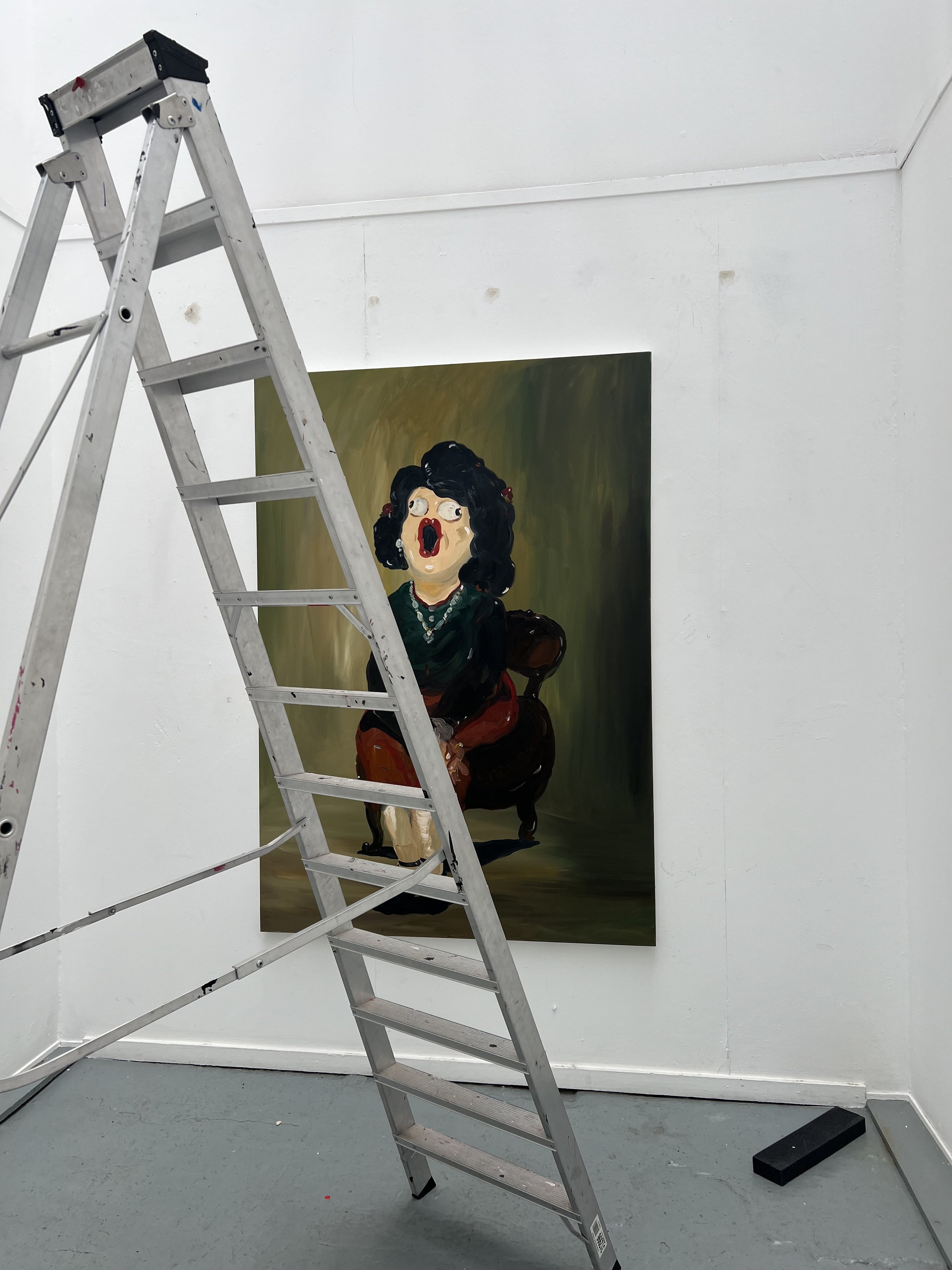 An art gallery room with a painting of a woman with black curly hair, wearing a black shirt and red pants, sitting on a chair with an expression of surprise or shock. An aluminum ladder is in the foreground, partially blocking the view of the painting.