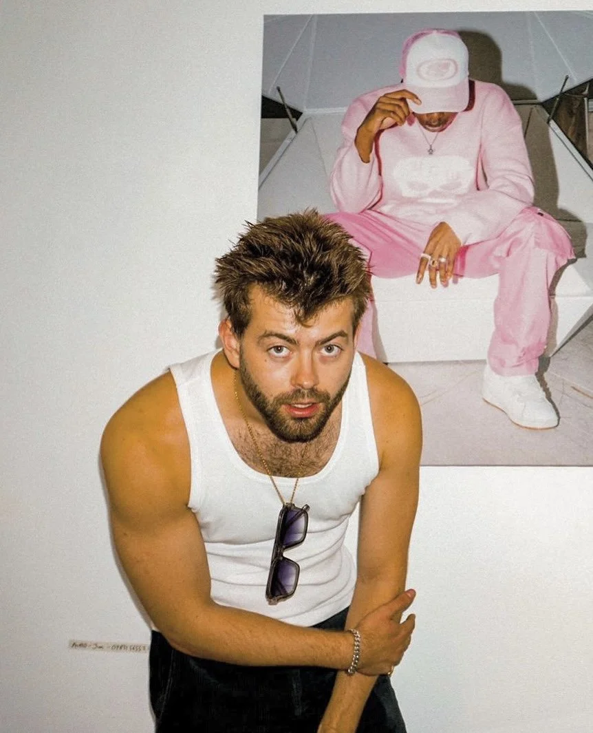 Man with brown hair and beard, wearing a white tank top, sunglasses hanging from his shirt, and a bracelet, standing in front of an artwork depicting a person in pink clothing and a white cap sitting on a chair.