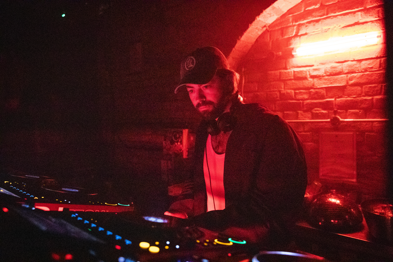 A DJ with headphones around his neck, wearing a baseball cap, working at a DJ booth in a dimly lit venue with red lighting and a brick wall background.