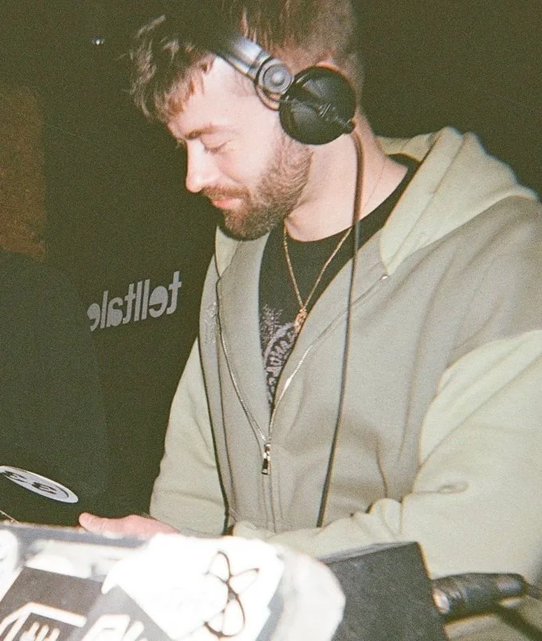 A DJ with headphones, wearing a beige hoodie, standing at a turntable in a dimly lit setting.