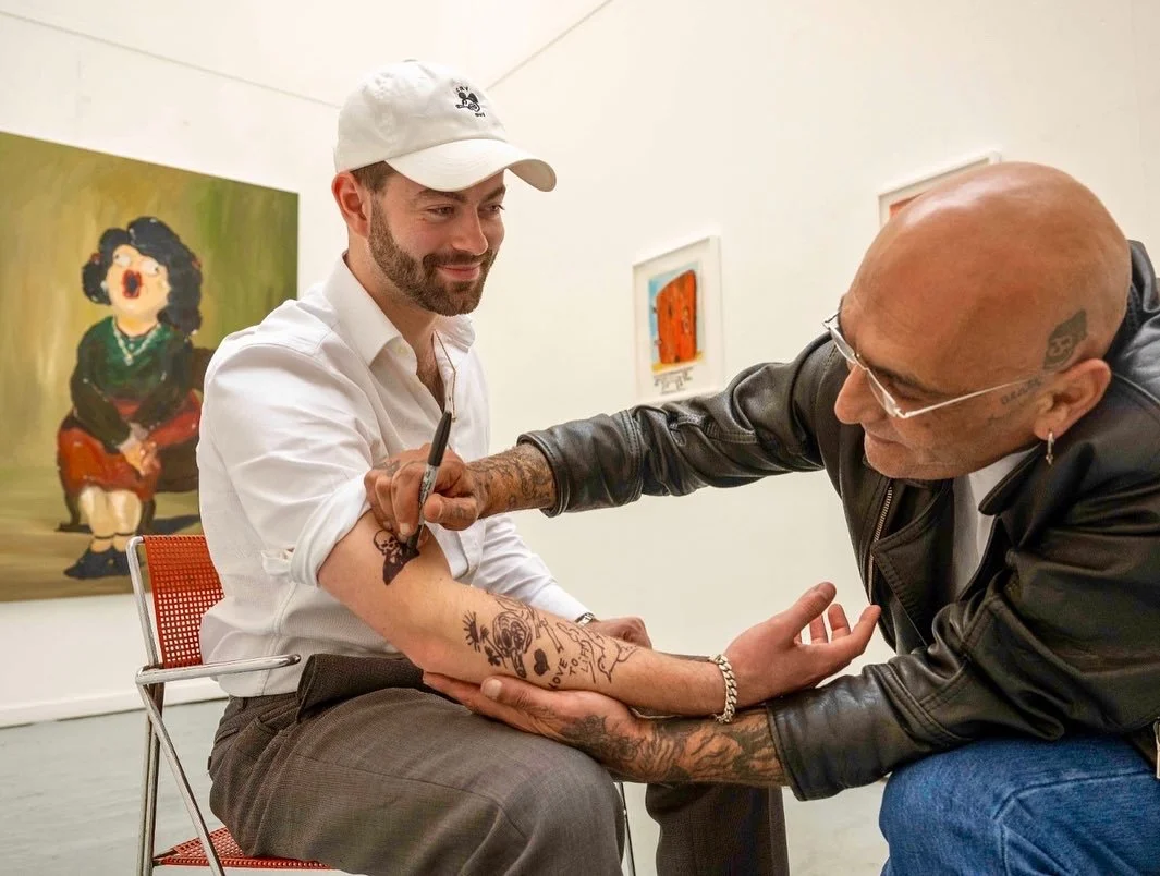 A man with a beard and tattoos getting his arm tattooed by an artist in an art gallery.