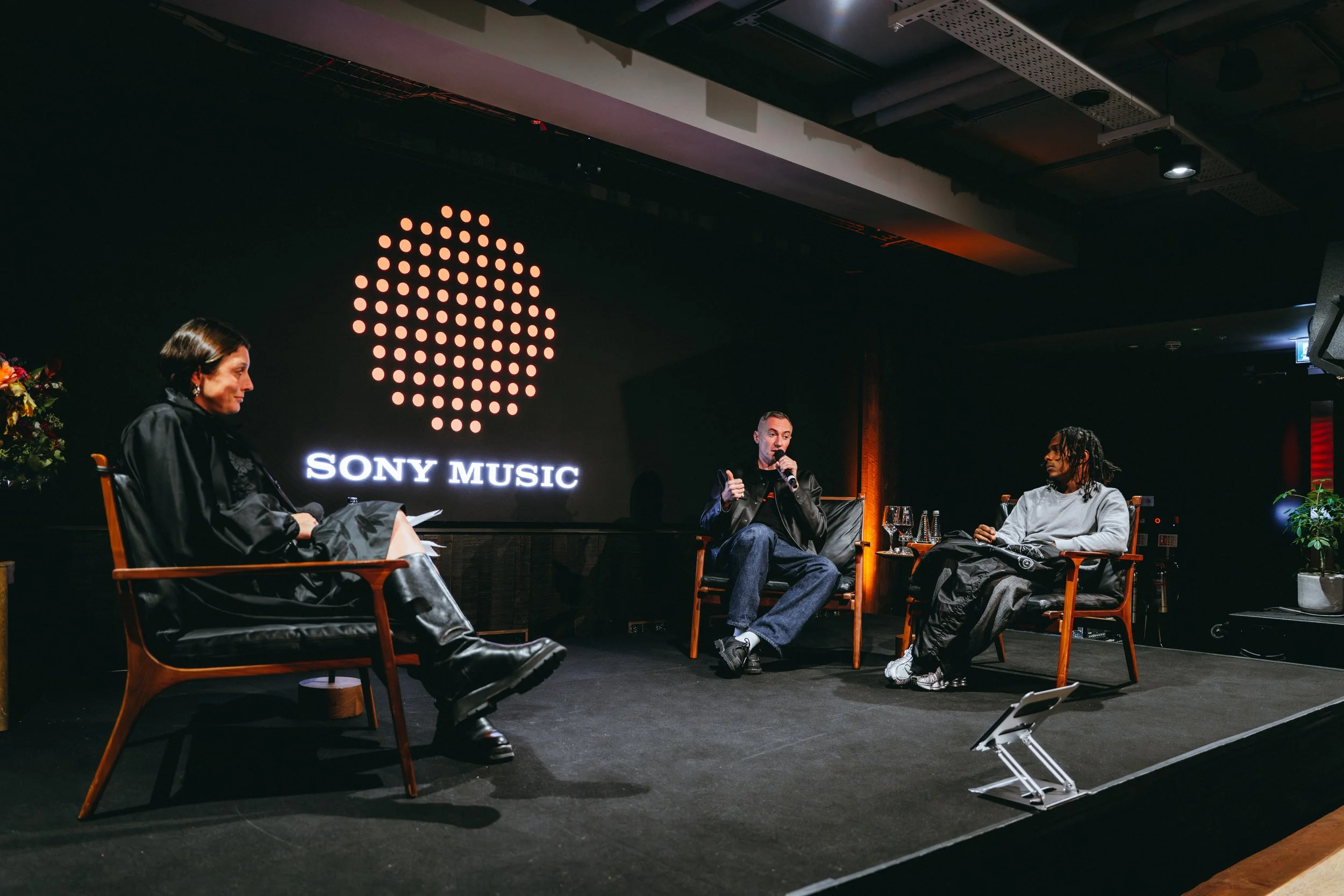 A panel discussion at Sony Music featuring three individuals seated on stage with a Sony Music logo displayed behind them. The person in the middle is speaking into a microphone, while the others listen.