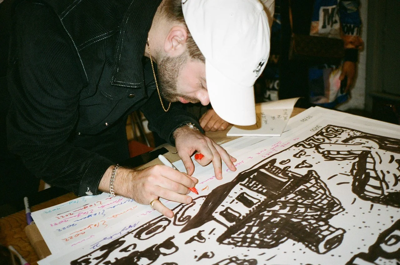 A man wearing a white cap and black clothing is drawing on a large sheet of paper with a black marker. The drawing appears to be a cartoonish house and other objects. There are multiple colored markers and handwritten notes around the drawing on the table.