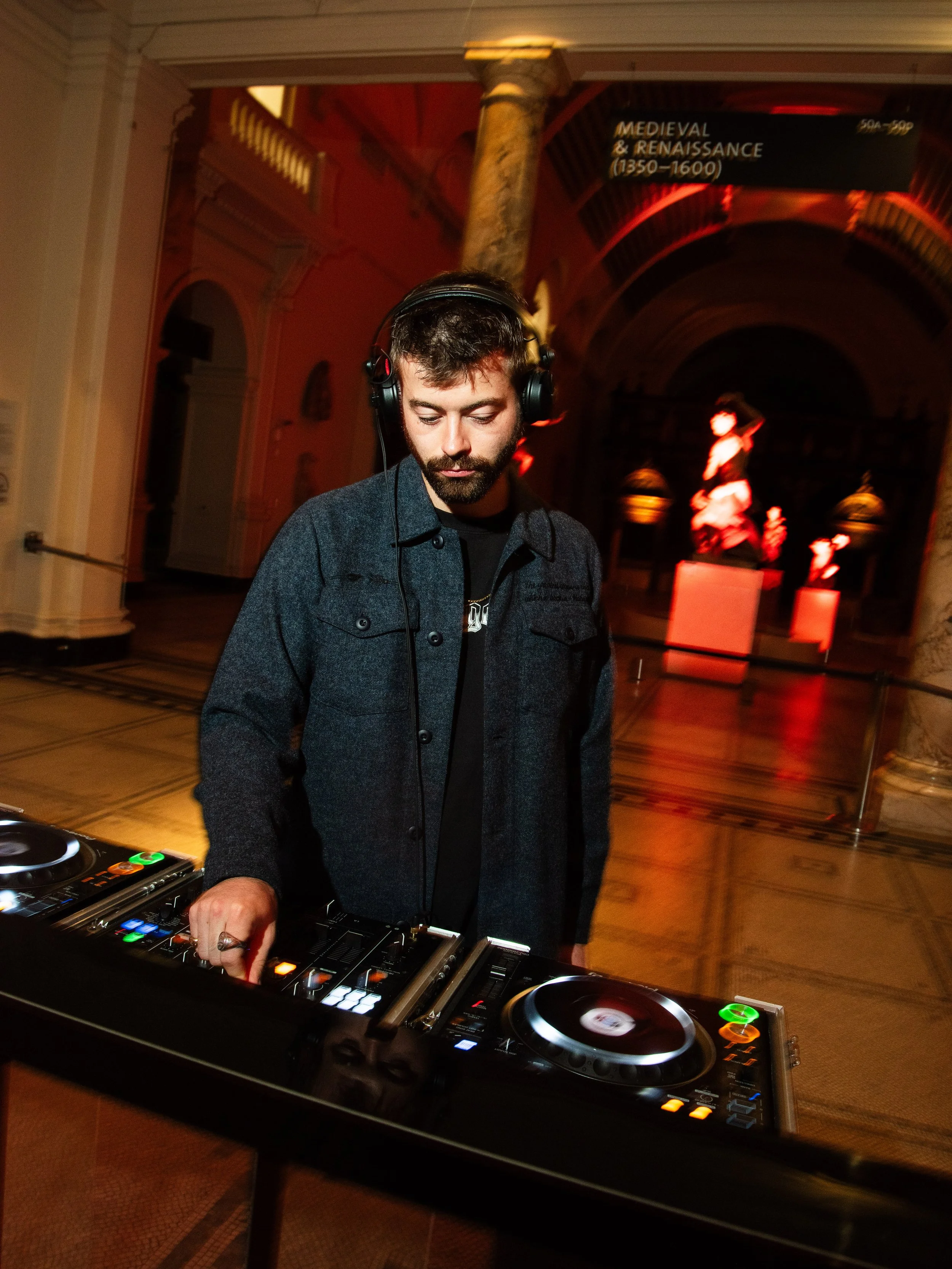 A DJ with headphones operating turntables in a historic venue with ornate architecture and sculptures, illuminated by red and orange lighting.