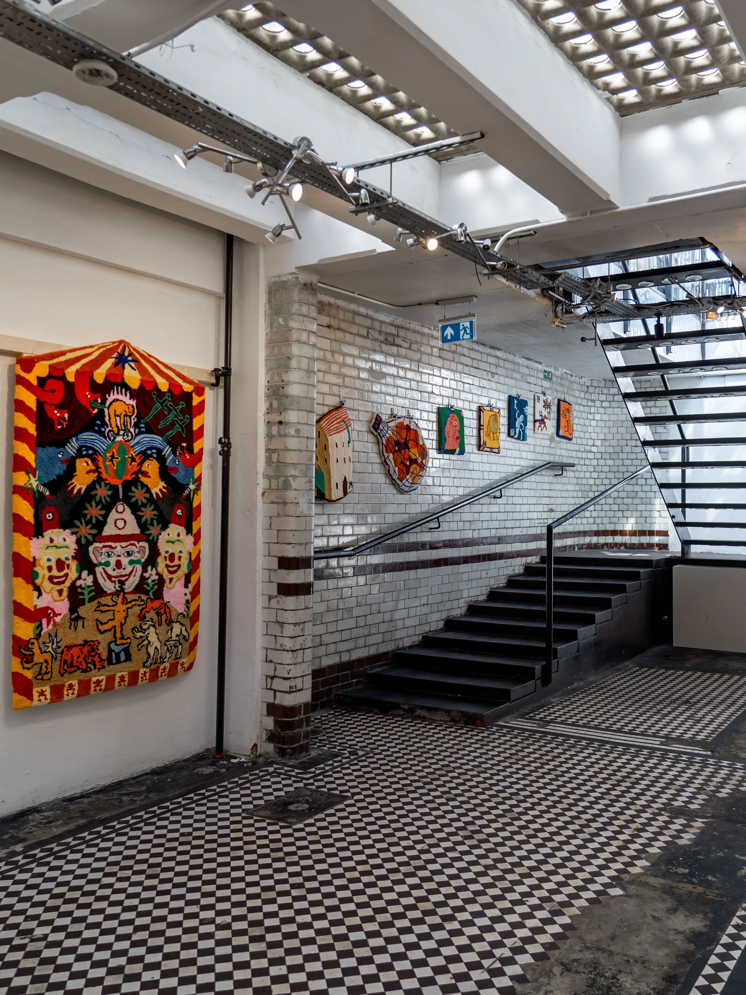 Art gallery interior with colorful textile art and smaller framed artworks on a white brick wall, black staircase, patterned tile floor, and ceiling with skylights.