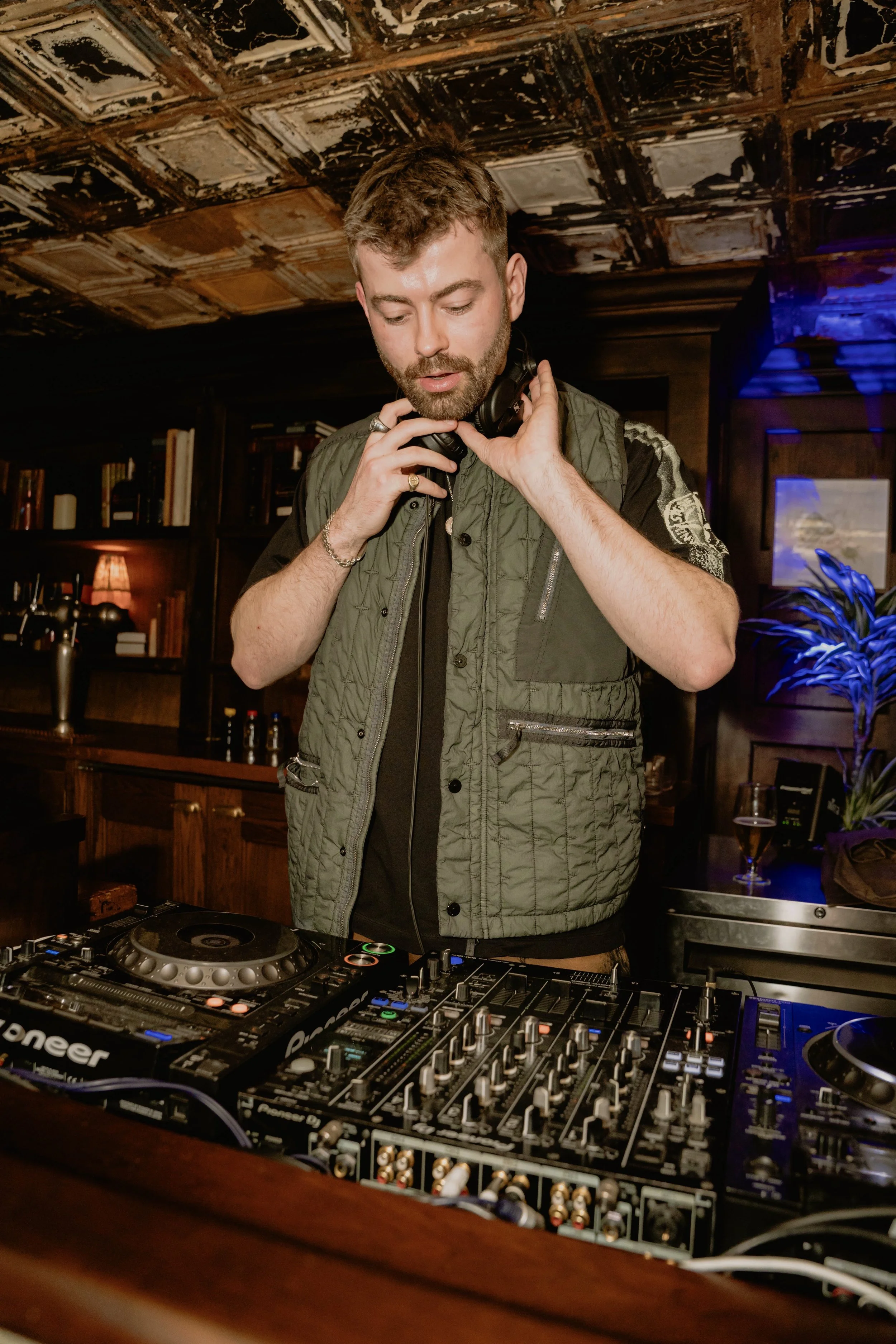 A man with a beard and short hair, wearing a green vest, is DJing with headphones. He is adjusting his headphones with one hand and standing behind a DJ controller in a dimly lit room.
