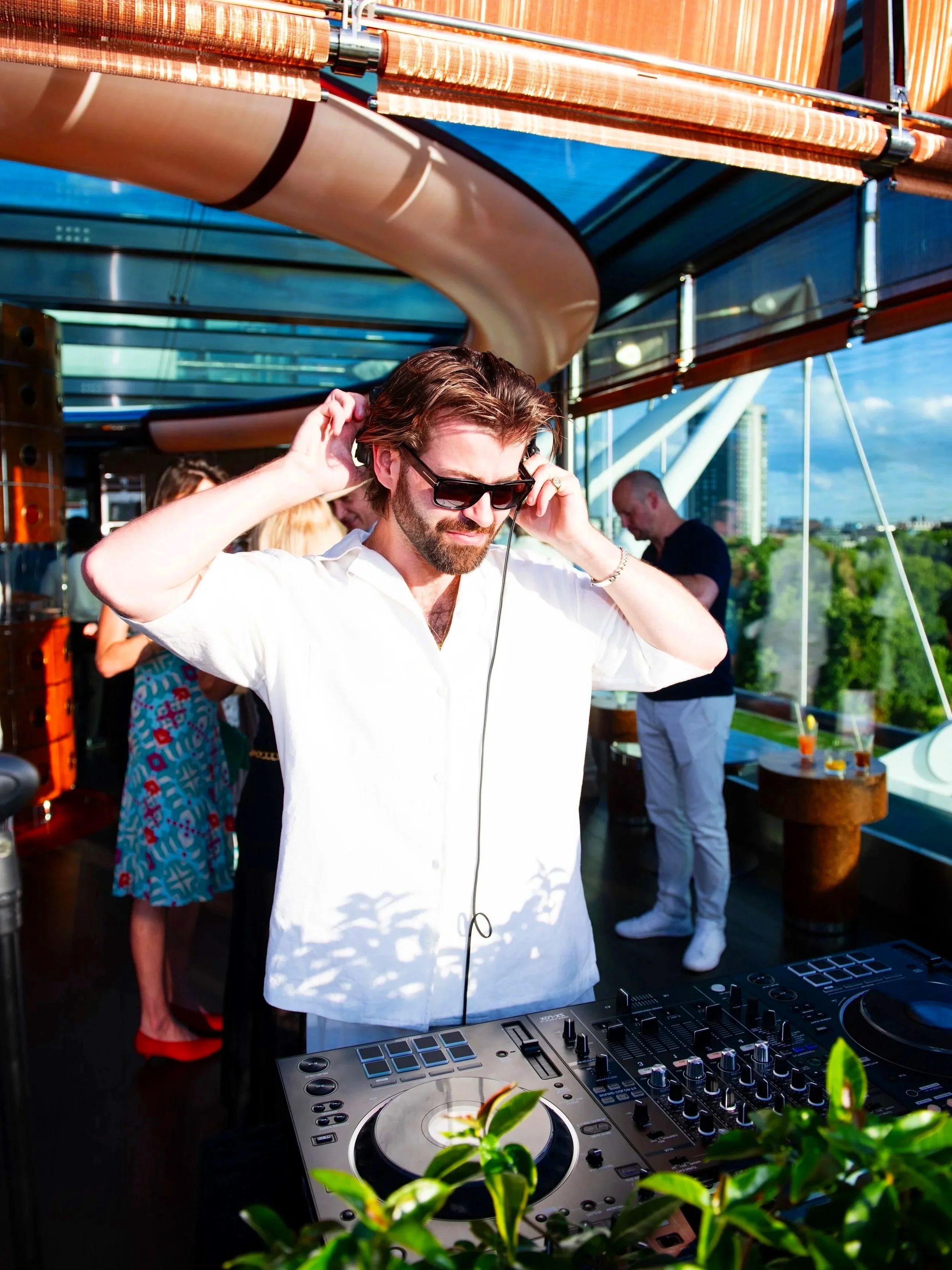 A man with sunglasses and a white shirt is DJing at an outdoor event on a rooftop, with a city skyline and green trees in the background.