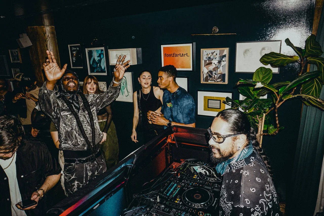 People dancing and enjoying music in a dark indoor venue with framed artwork on the wall and a DJ playing music.