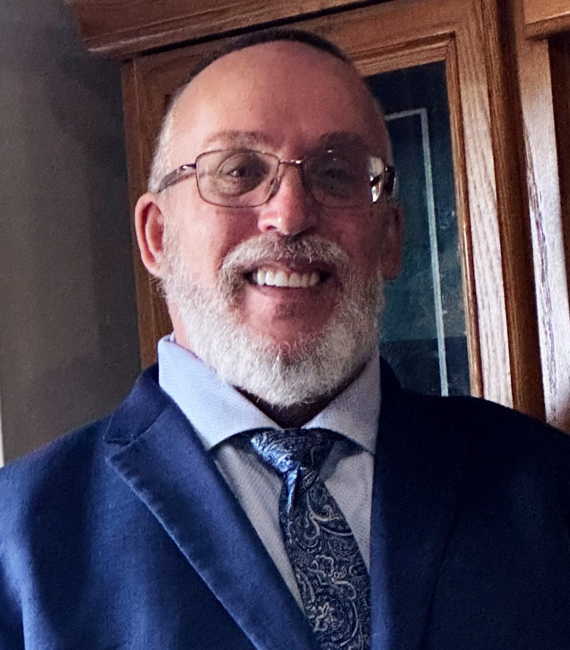 A smiling man with glasses, a gray beard, wearing a navy blue suit, light blue shirt, and patterned tie, standing in front of a wooden cabinet.