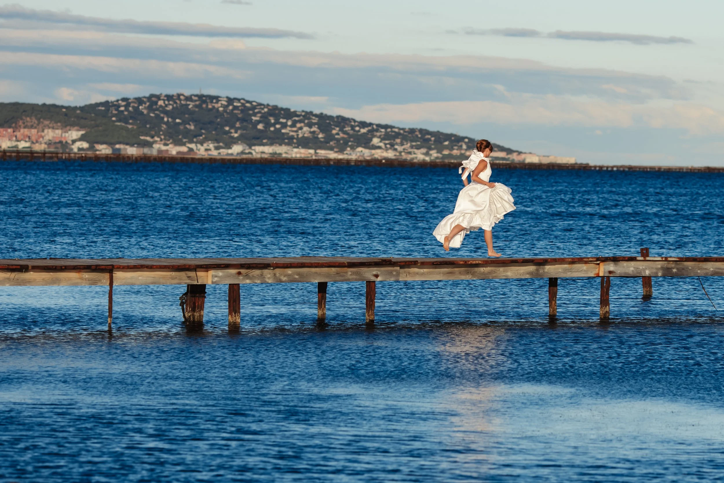 Femme en robe blanche courant sur une jetée en bois au bord de l'eau, avec une ville et une colline en arrière-plan.