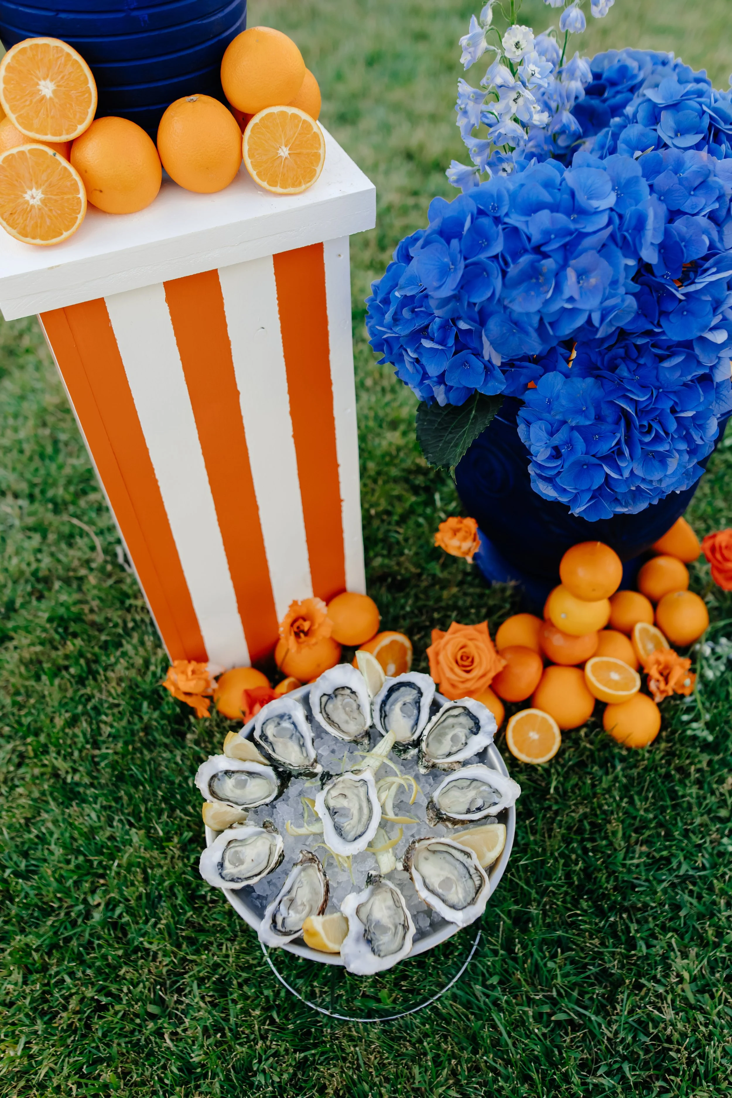Plate d'huîtres avec du citron, décorée d'oranges et de roses, à côté d'un vase de fleurs bleues, avec des oranges et des roses orange sur le sol