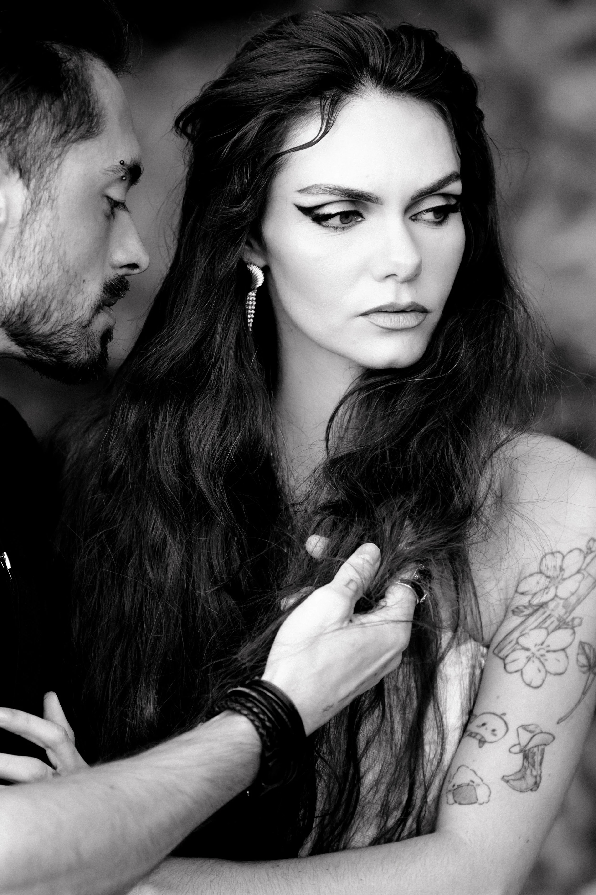 Une femme aux longs cheveux foncés, porteur de boucles d'oreilles, regardant à droite. Un homme prend sa chevelure en main, près de son épaule tatouée de fleurs, champignons et autres motifs.