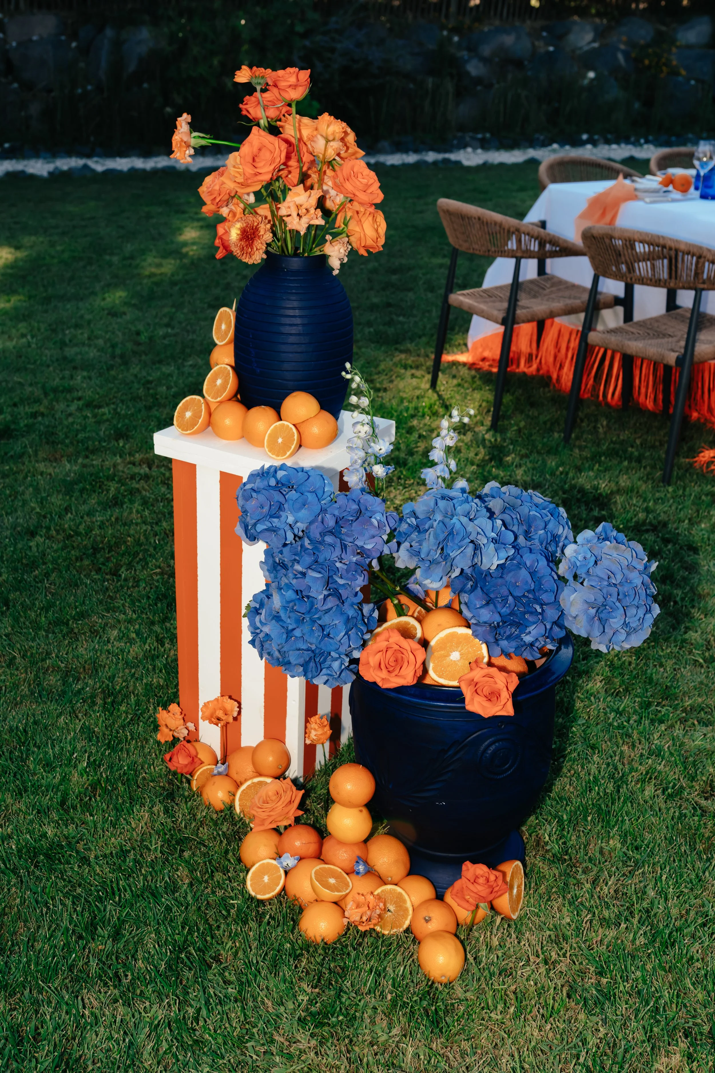 Une décoration de fête avec des vases bleus contenant des fleurs orange et bleues, accompagnés de tranches d'orange, sur une table et le sol, dans un jardin avec tables et chaises en arrière-plan.