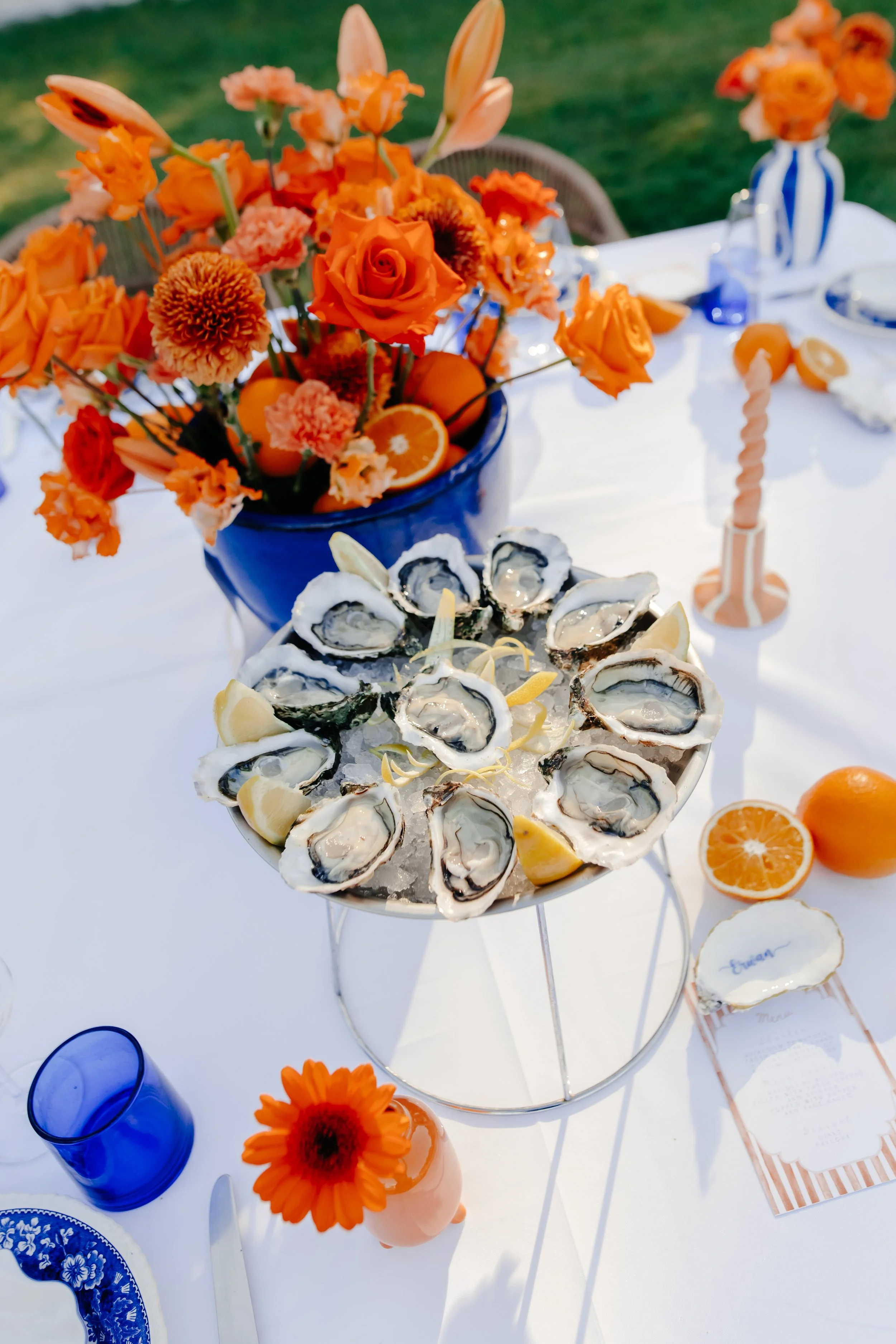 Plate d'huîtres sur glace avec des quartiers de citron, au centre une grande vase avec des fleurs orange, une table dressée avec une bougie torsadée rose, une petite vase avec une fleur orange, et des verres bleus, en extérieur.