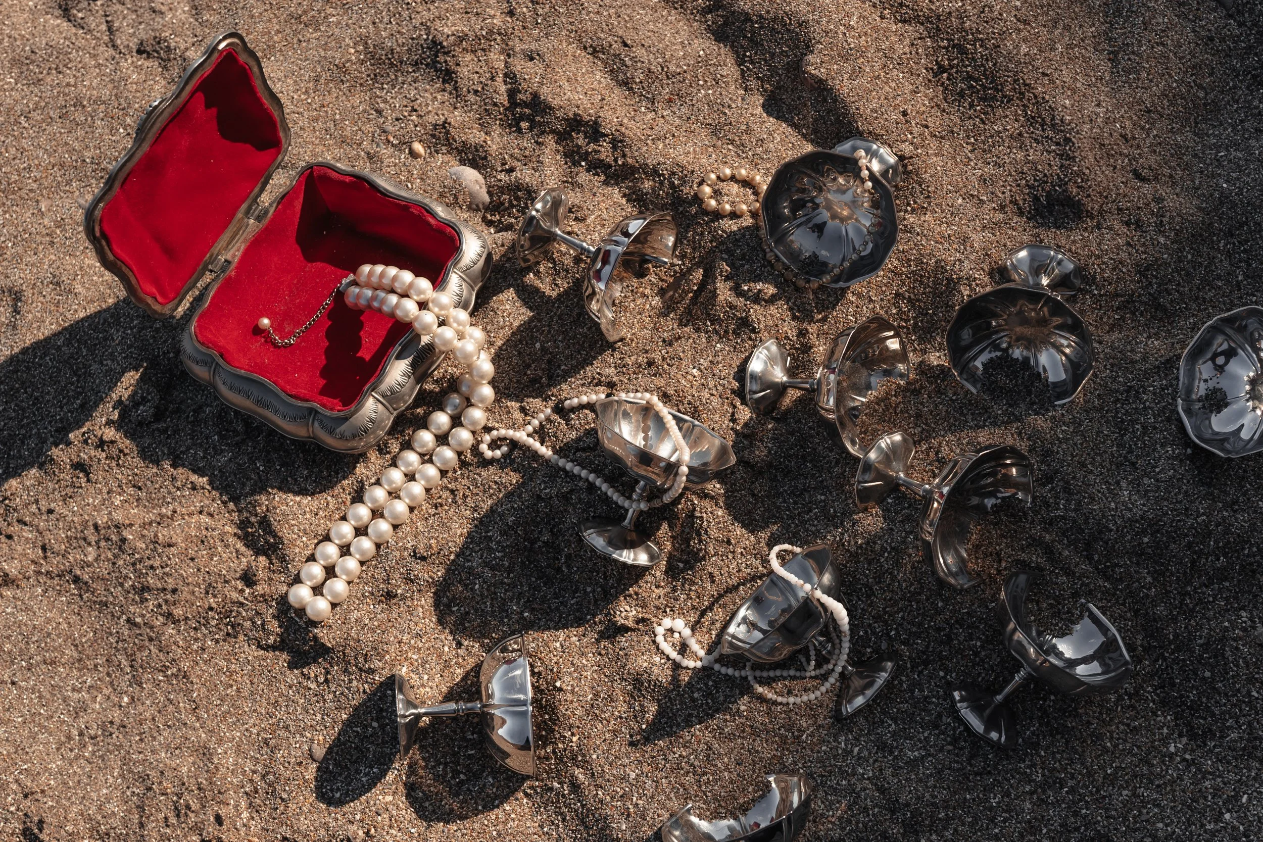Bijoux et verres en argent dispersés sur le sable avec une boîte à bijoux en velours rouge