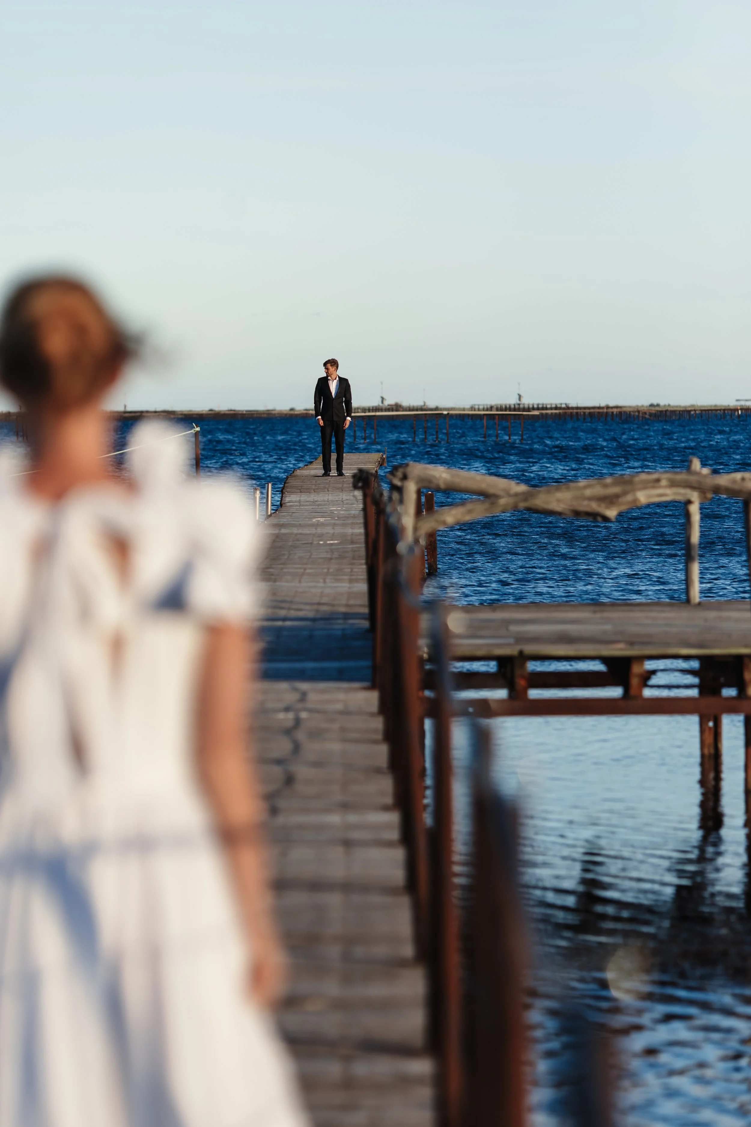 Une femme en robe blanche, floue au premier plan, regarde un homme en costume noir à l'arrière-plan, au bout d'une jetée en bois sur la mer.