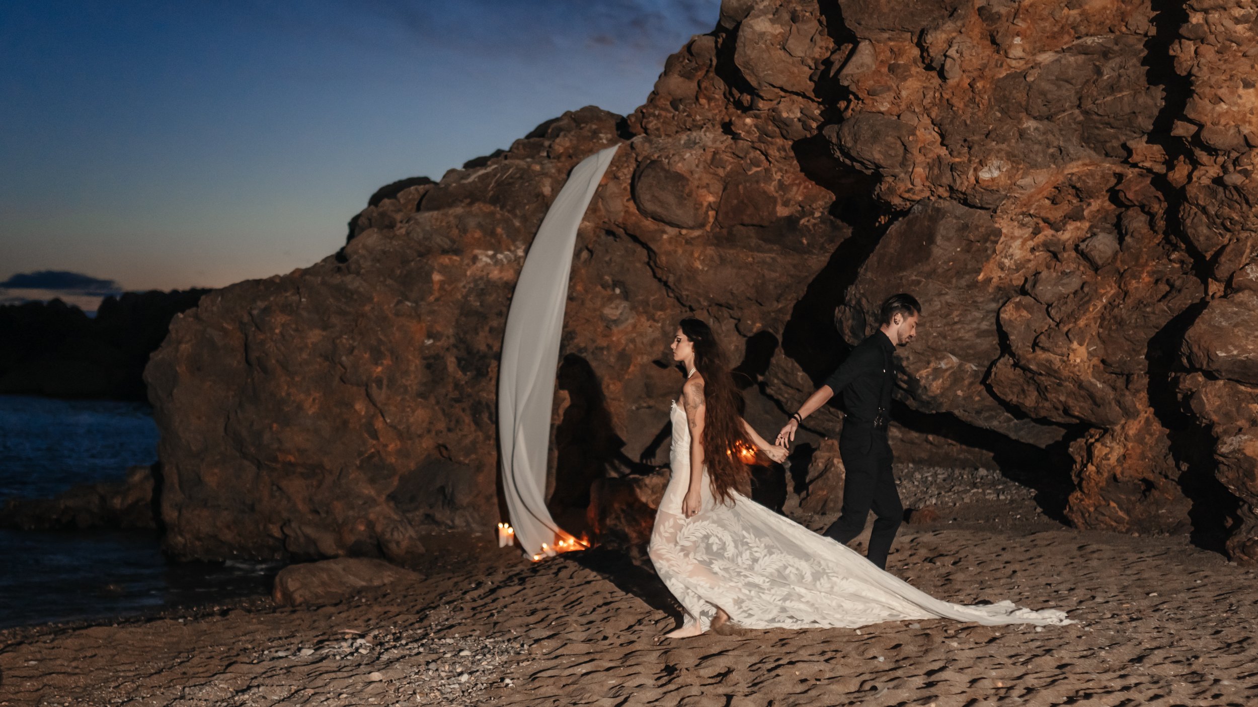 Un couple marchant sur la plage au coucher du soleil, la femme porte une robe de mariée longue et le homme un costume noir.