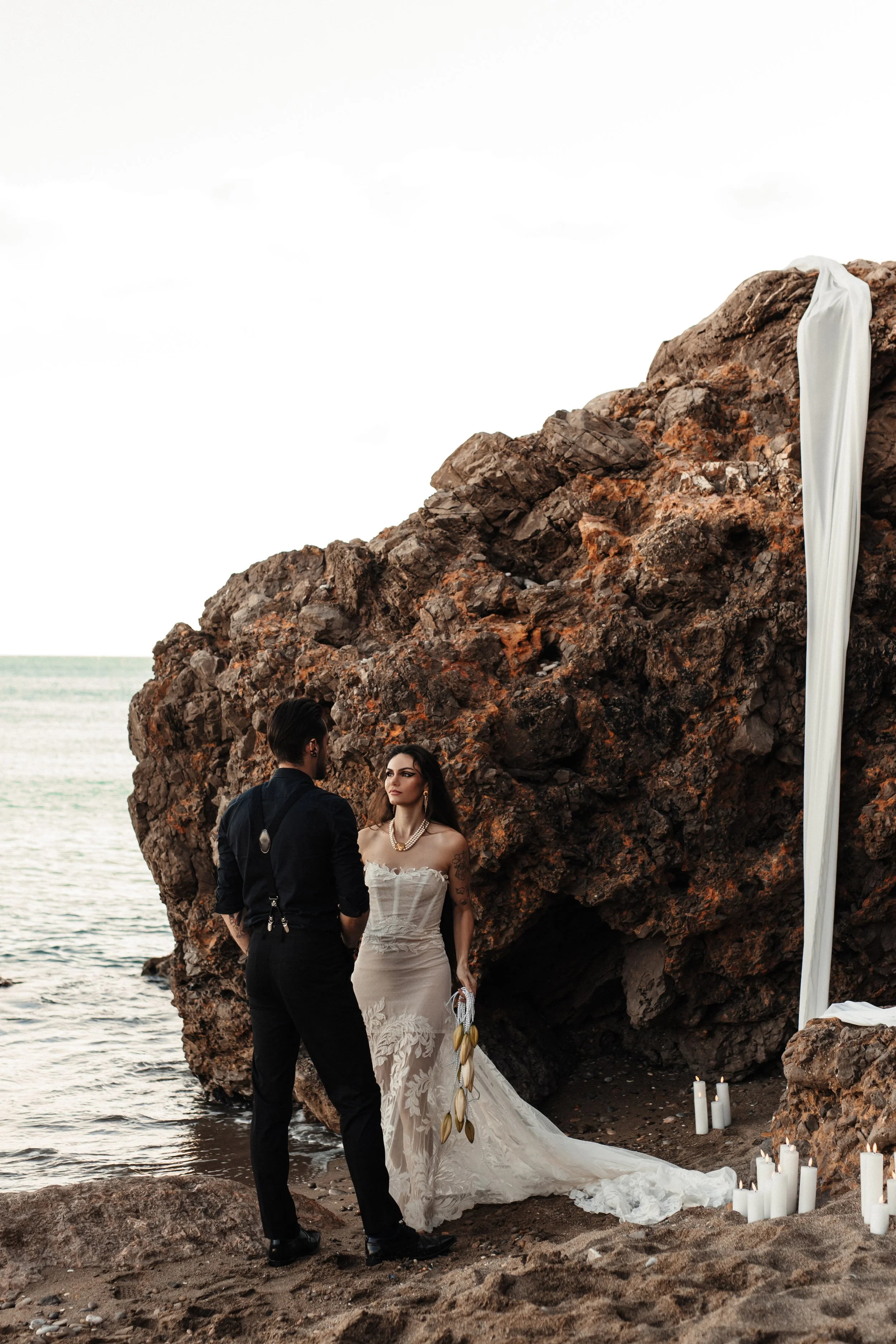 Un couple se tenant devant une scène de mariage sur la plage, avec des rochers en arrière-plan, des bougies et un drap blanc accrochés à la roche.