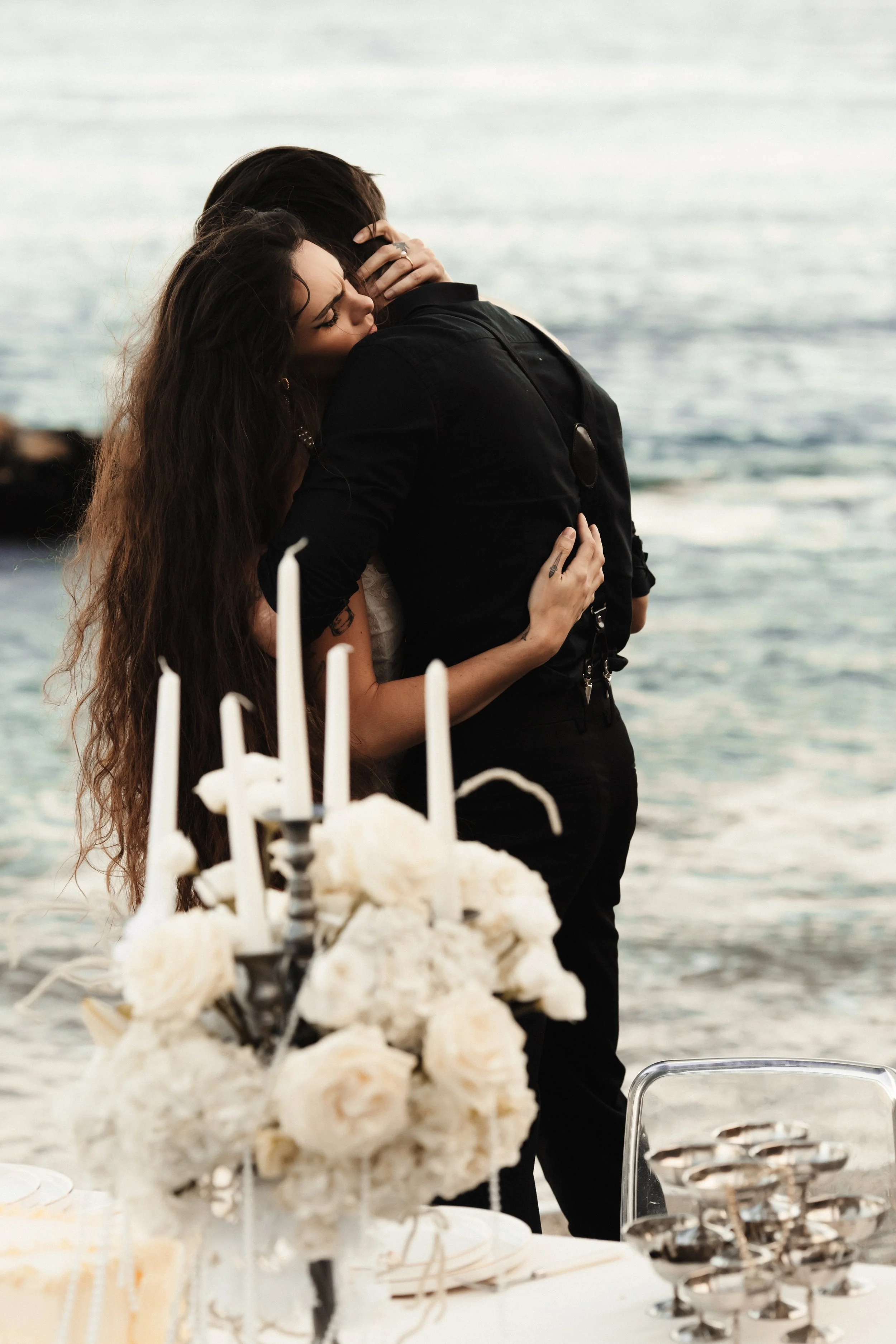 Deux personnes s'embrassent sur une plage lors d'une fête en plein air avec des fleurs blanches et des bougies sur la table.