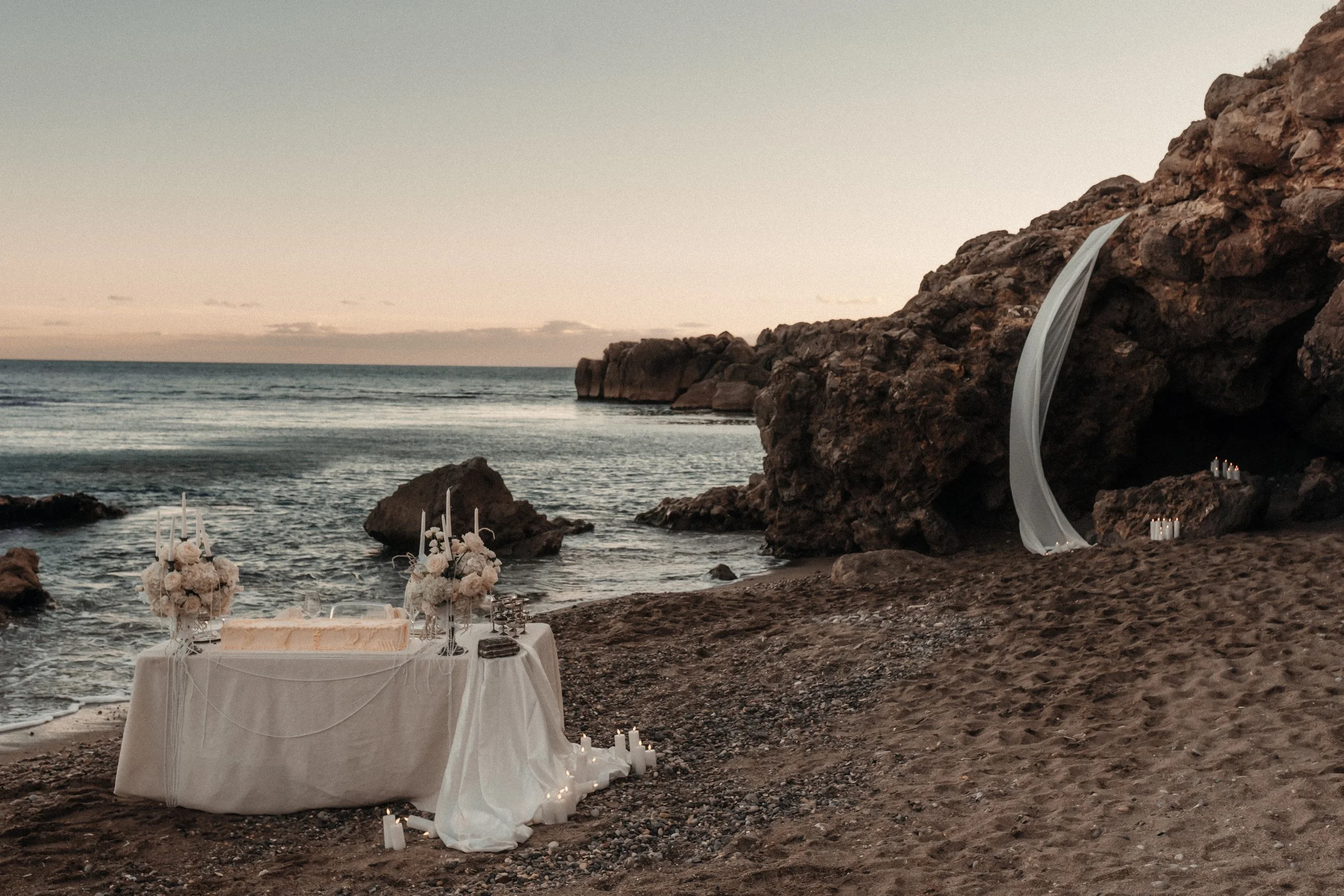 Décor de mariage romantique sur la plage avec un altar rocheux, voilage blanc, table élégante avec fleurs blanches et bougies, crépuscule.