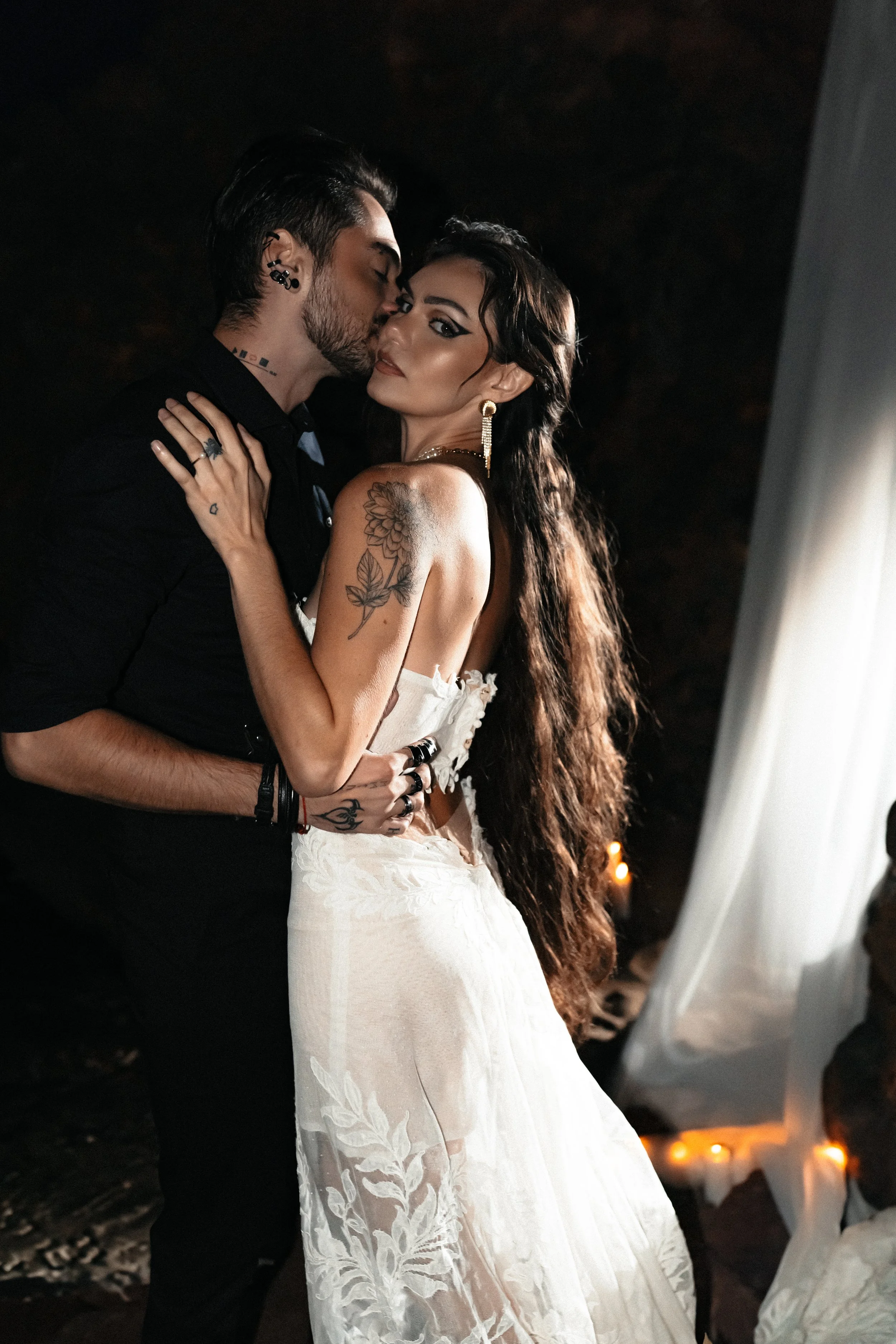 Un couple danse lors d'une cérémonie à l'intérieur, la femme porte une robe blanche en dentelle et a une tatouage sur le bras, l'homme porte une chemise noire.