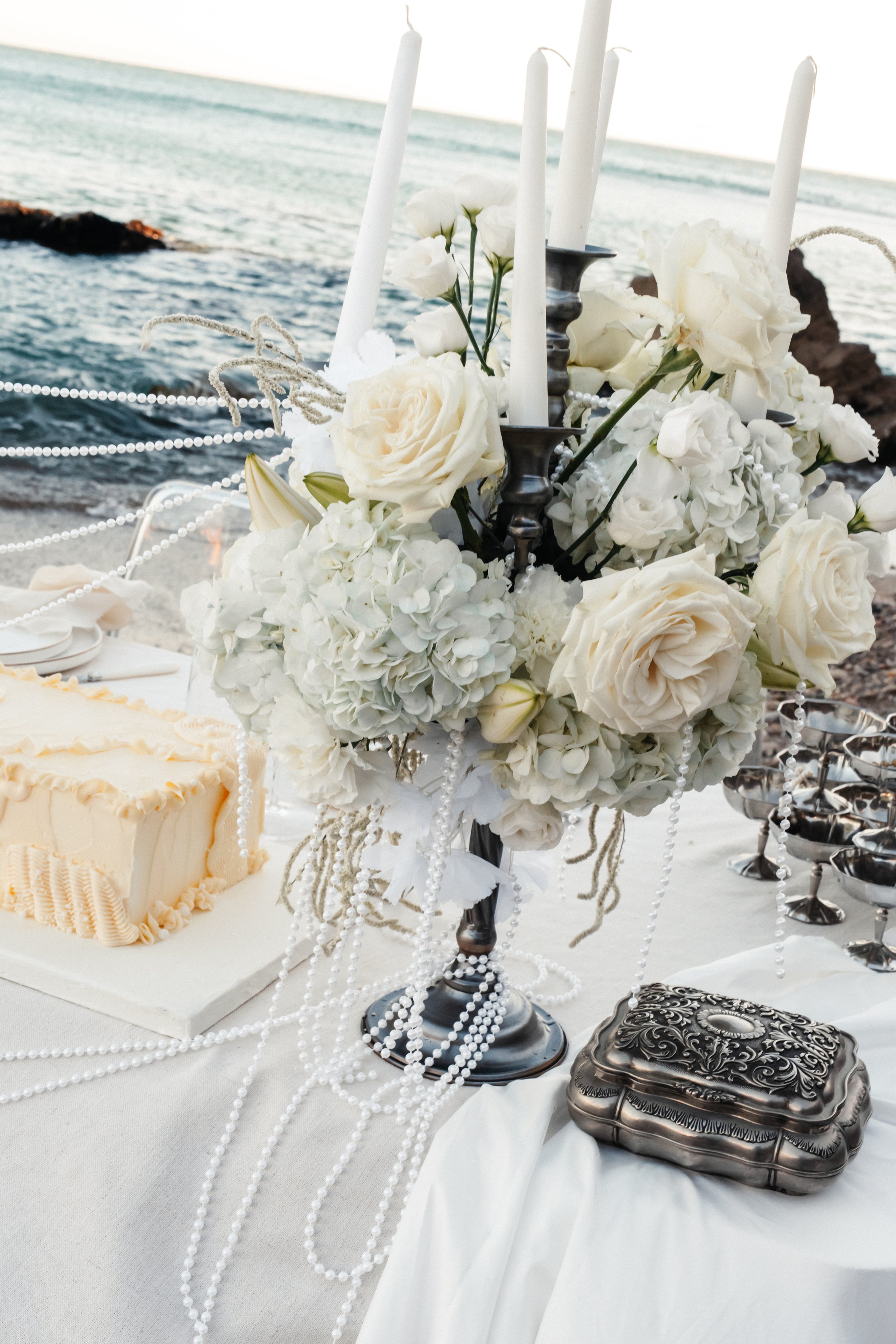 Décoration de mariage avec bouquet de fleurs blanches, chandeliers, et gâteau sur une table près de la mer.