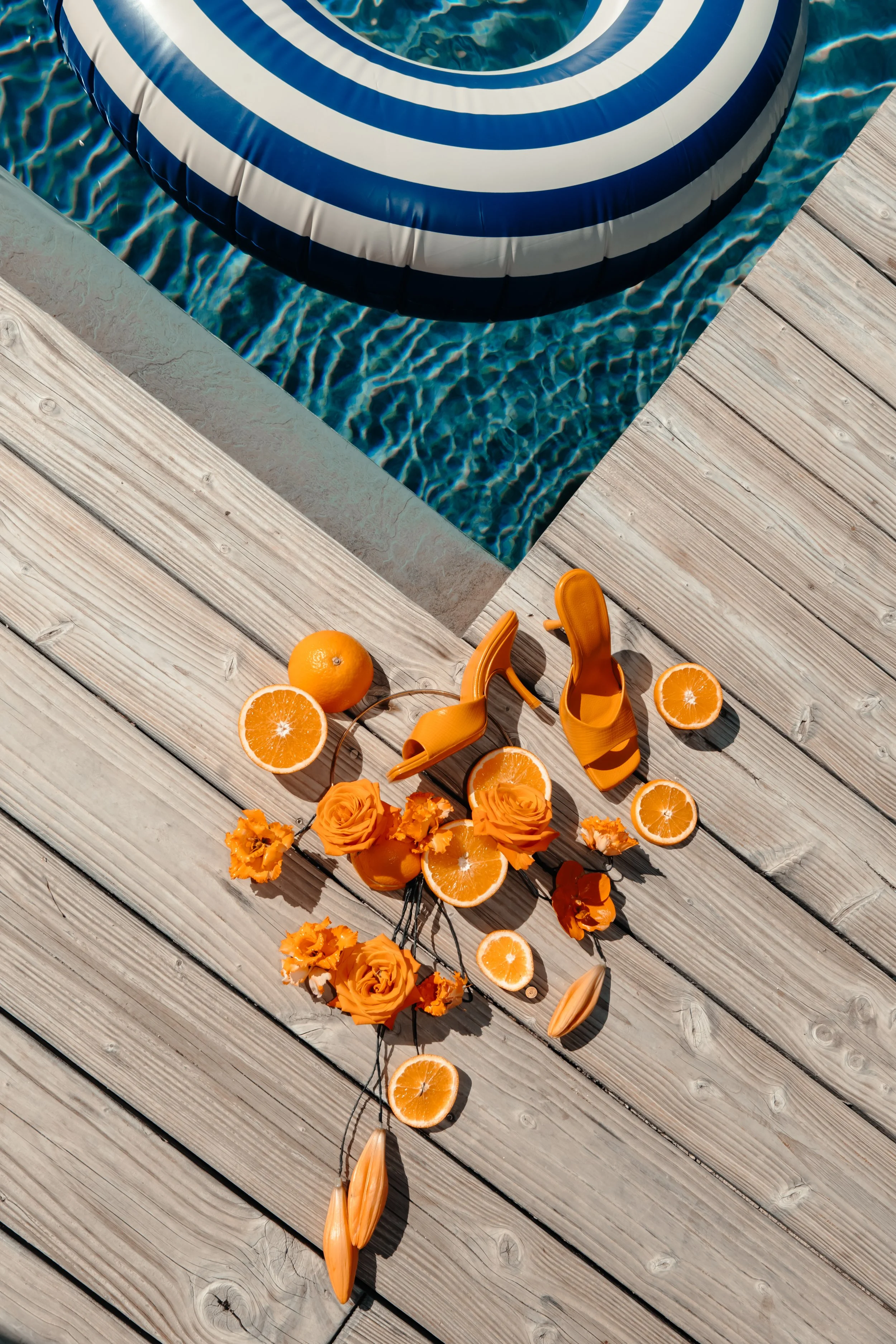 Décoration estivale avec des fleurs oranges, des tranches d'orange, des sandales orange, un cerise et un parasol à la piscine.
