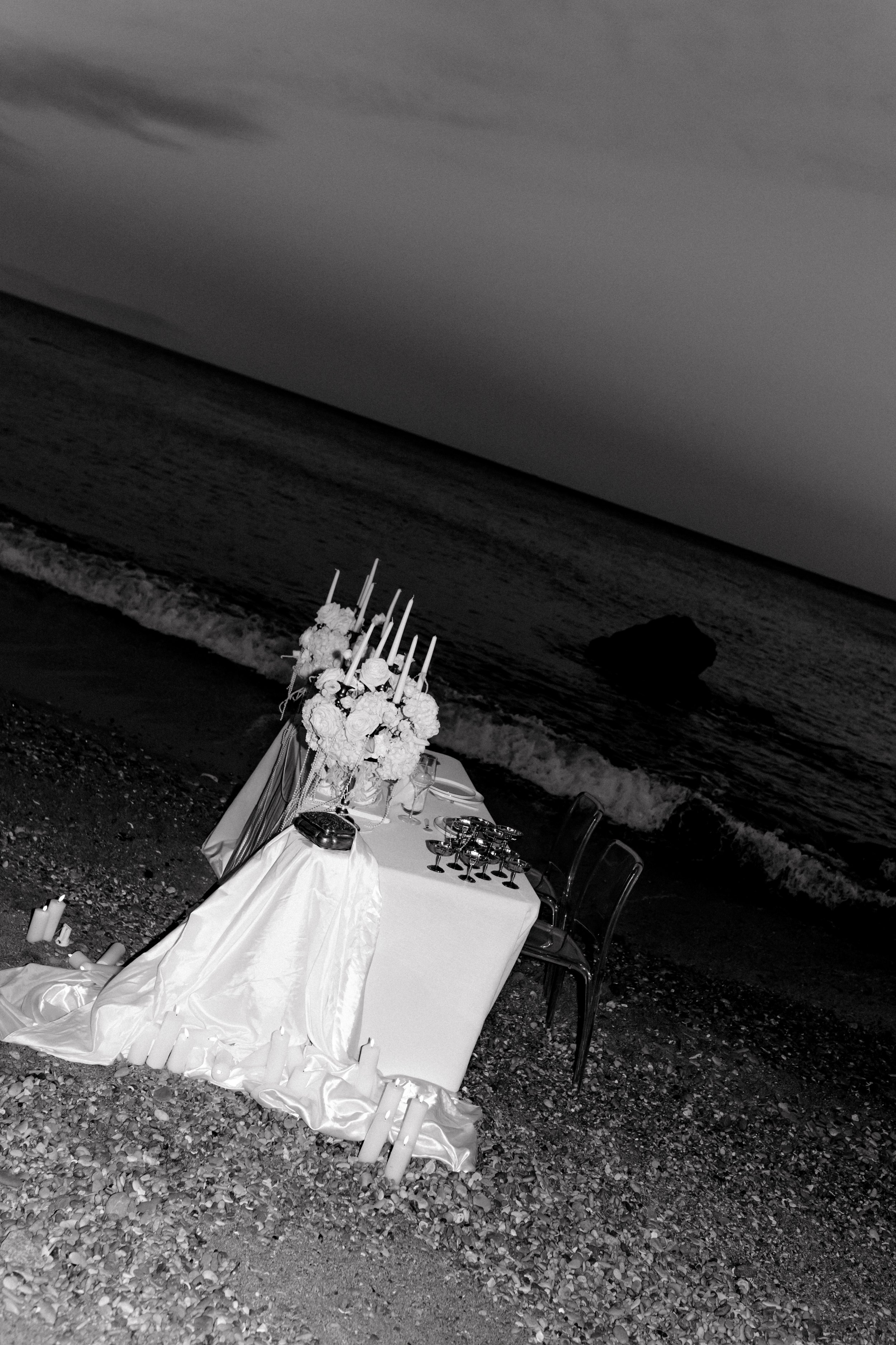 Une table décorée avec des fleurs, des chandelles et des verres, située sur une plage au bord de la mer, à la tombée de la nuit.