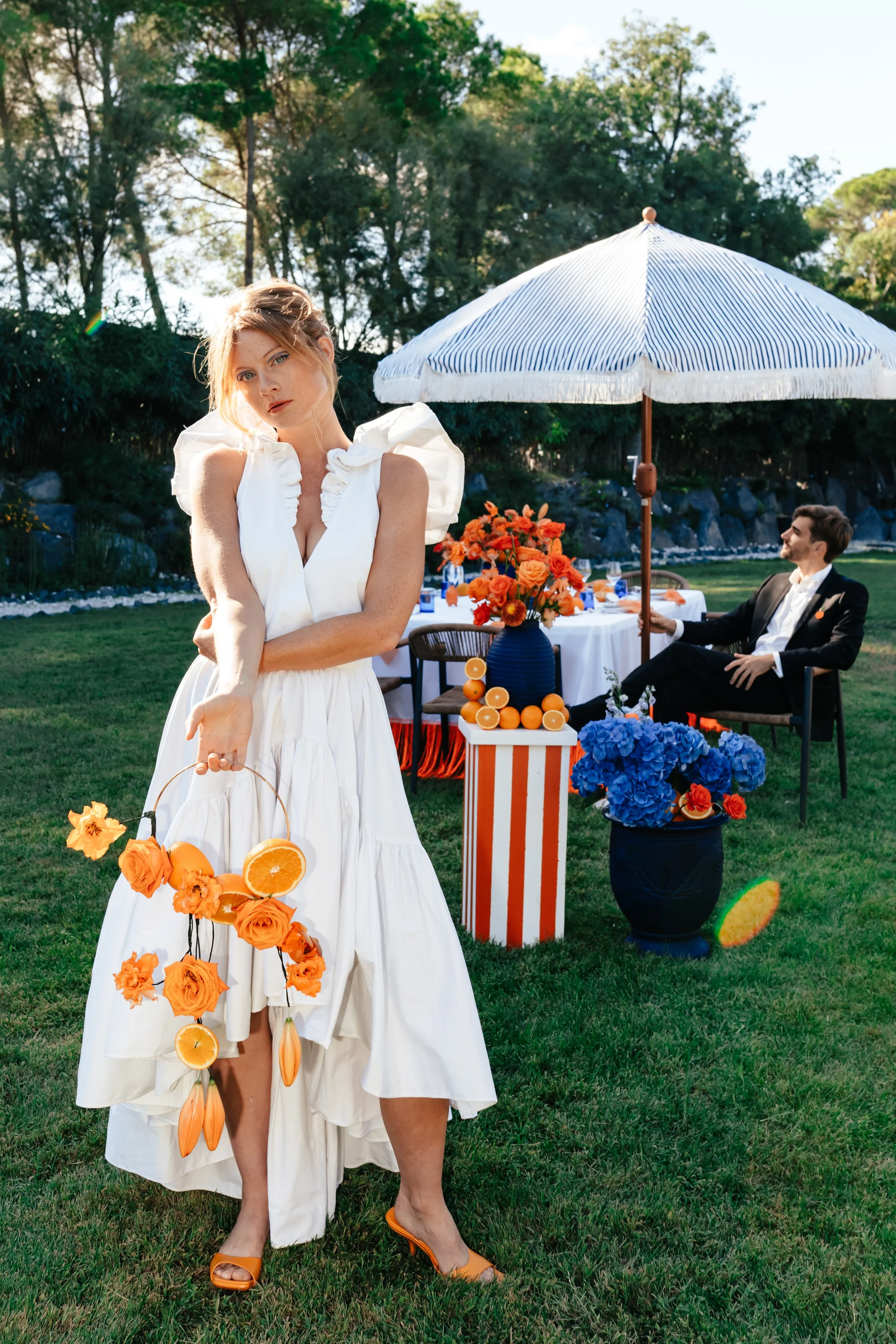 Une femme en robe blanche avec des accessoires d'orange, tenant un panier de fleurs d'orange, se tient devant une table d'événement en plein air, avec une manique blanche et un homme en costume sombre assis sous un parasol à l'arrière-plan.