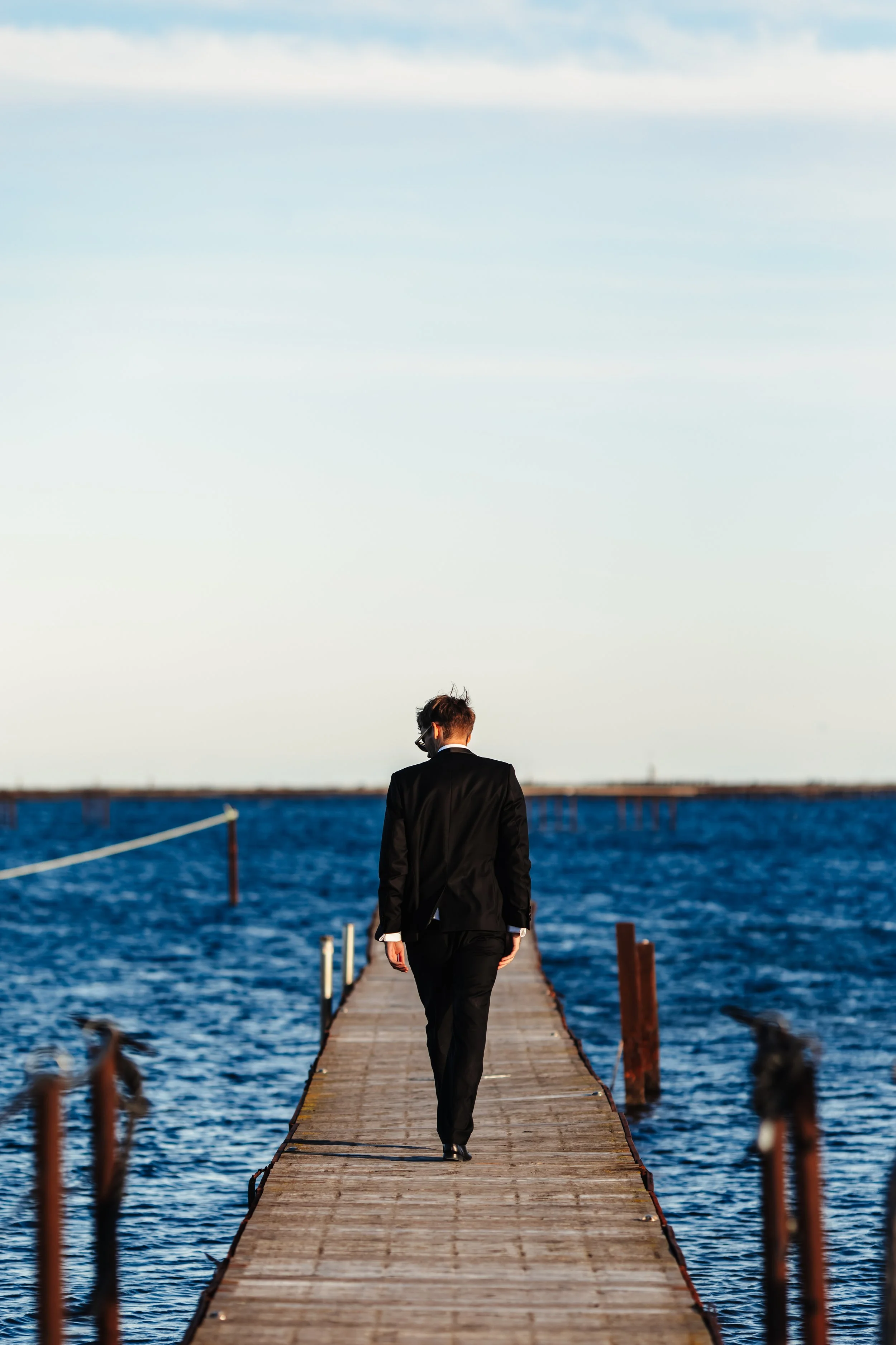 Un homme en costume noir marche seul sur un quai en bois au bord de l'eau, avec la mer et un ciel clair en arrière-plan.
