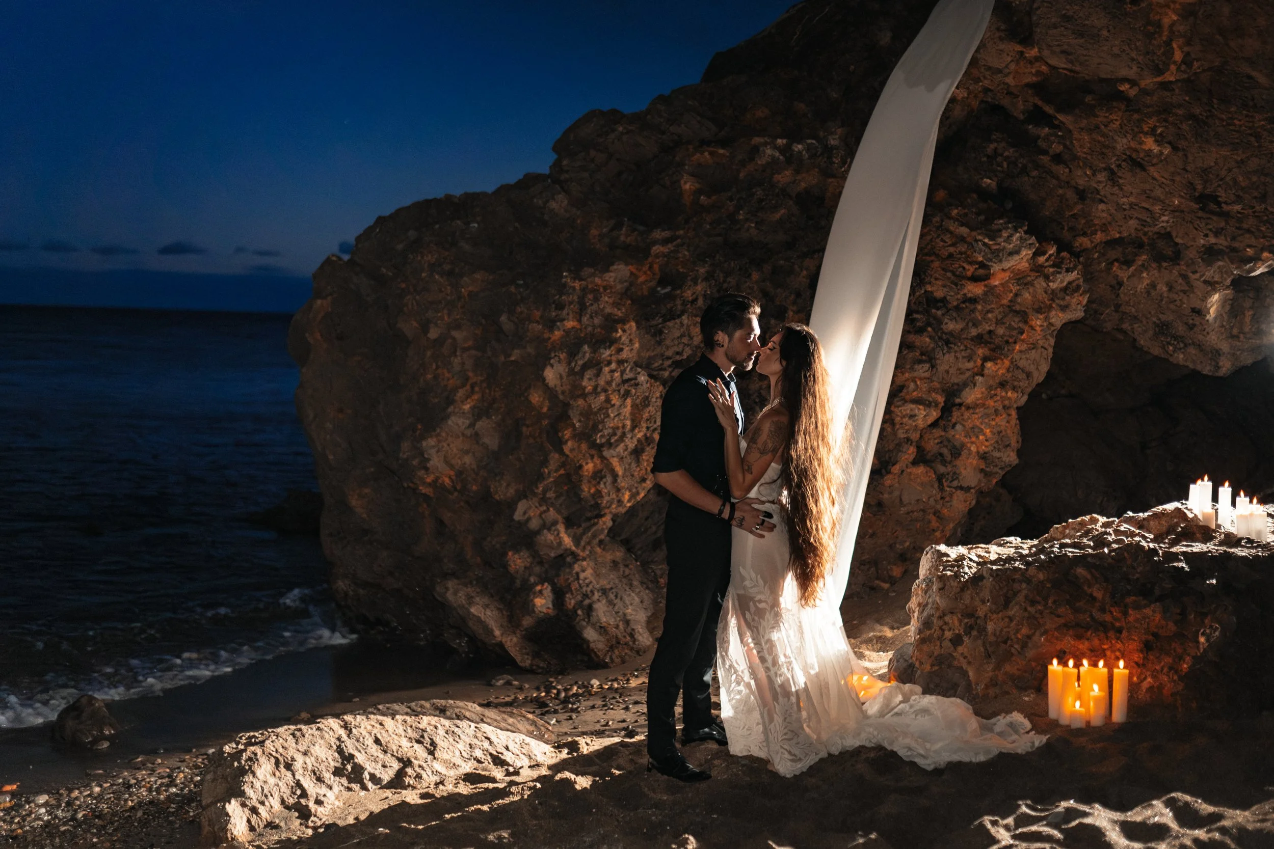 Un couple se tenant proche, dans une ambiance romantique, sur une plage rocheuse avec des bougies allumées, à la tombée de la nuit.