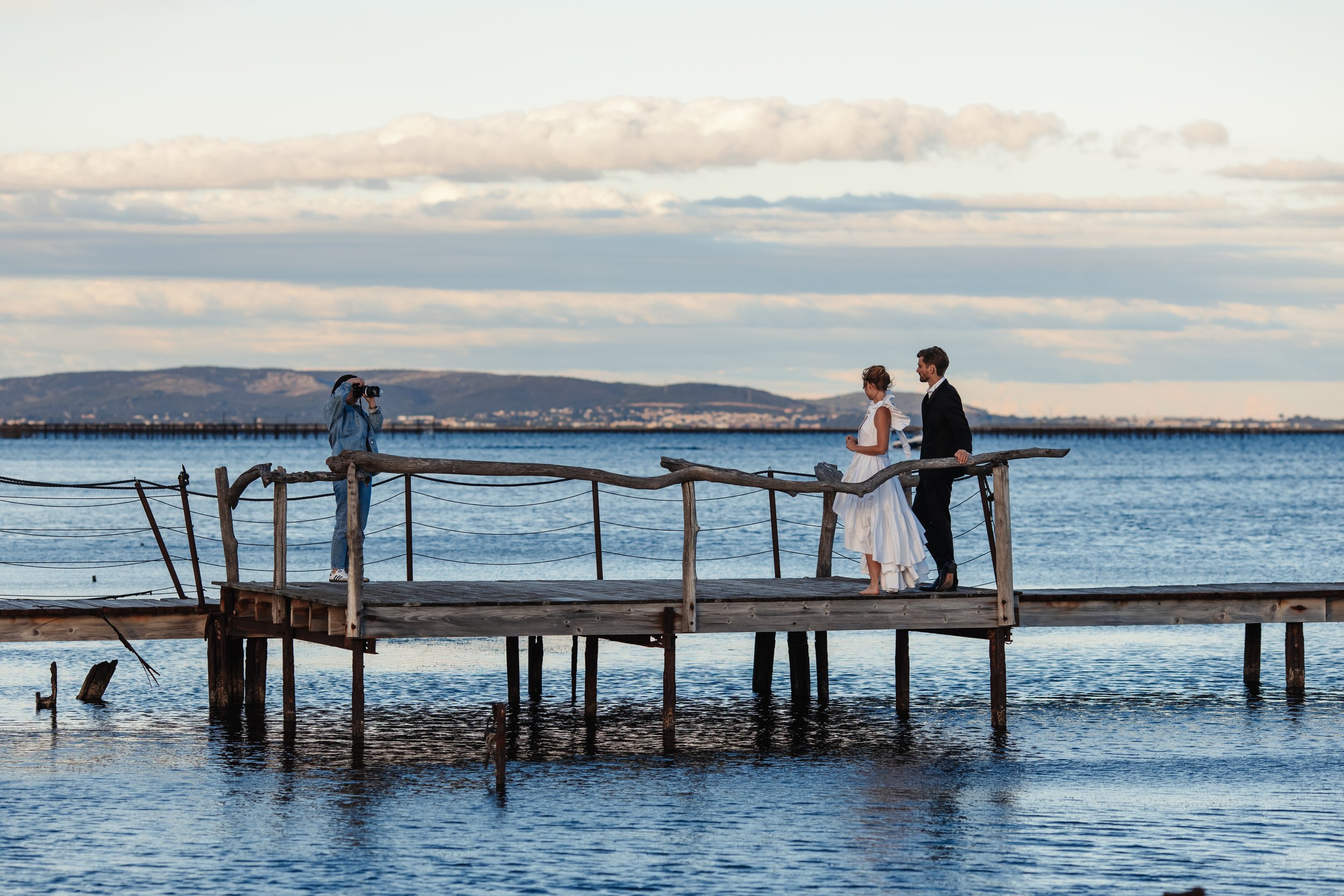 Un couple habillé en tenue de mariage se tient sur une jetée en bois au bord de l'eau, avec une photographe les photographiant, contre un ciel orageux.