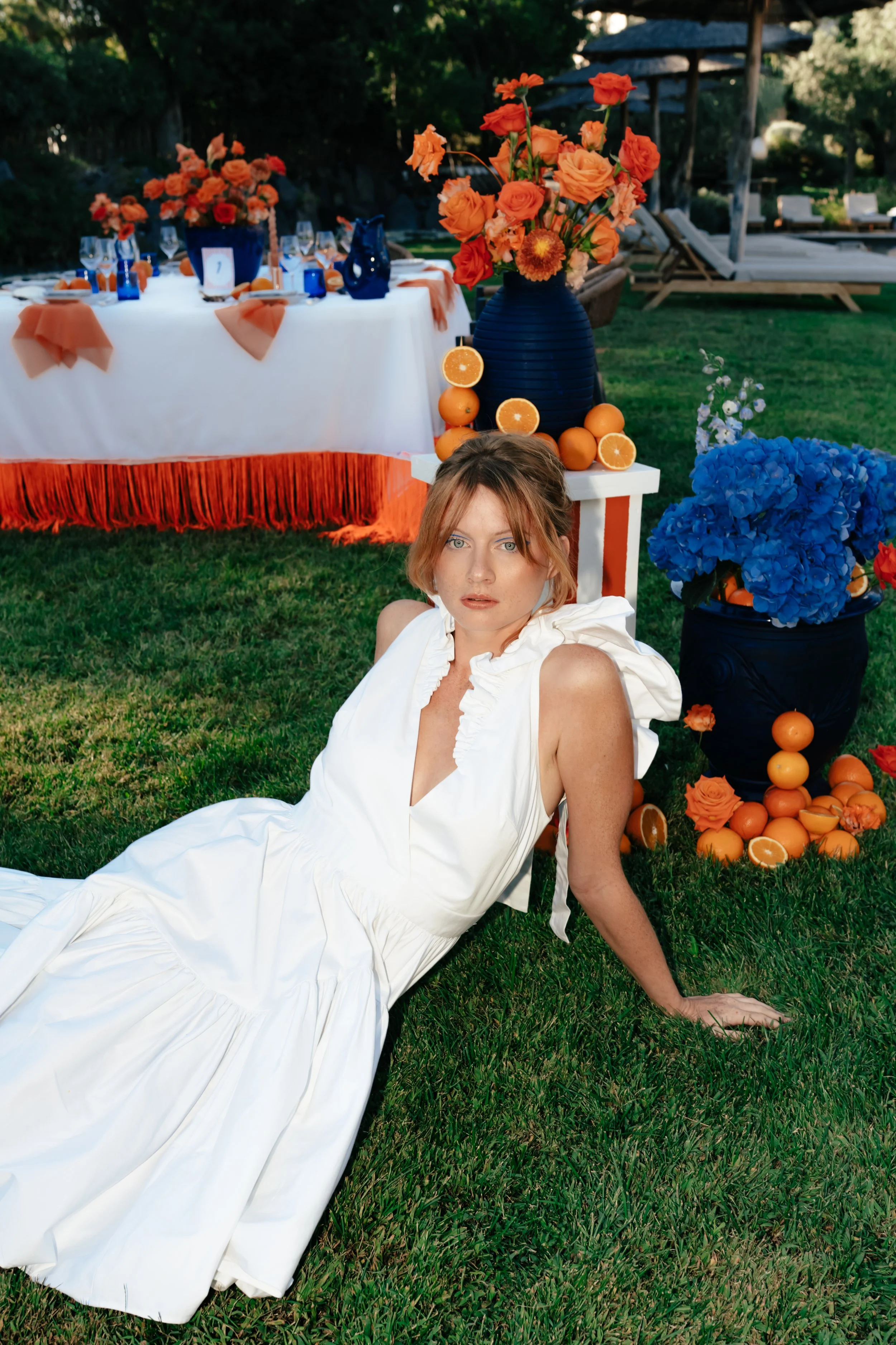Jeune femme en robe blanche allongée sur l'herbe devant une décoration de fête avec des vases contenant des fleurs orange et bleues, des oranges coupées en morceau, et une table en arrière-plan décorée de tissus orange.