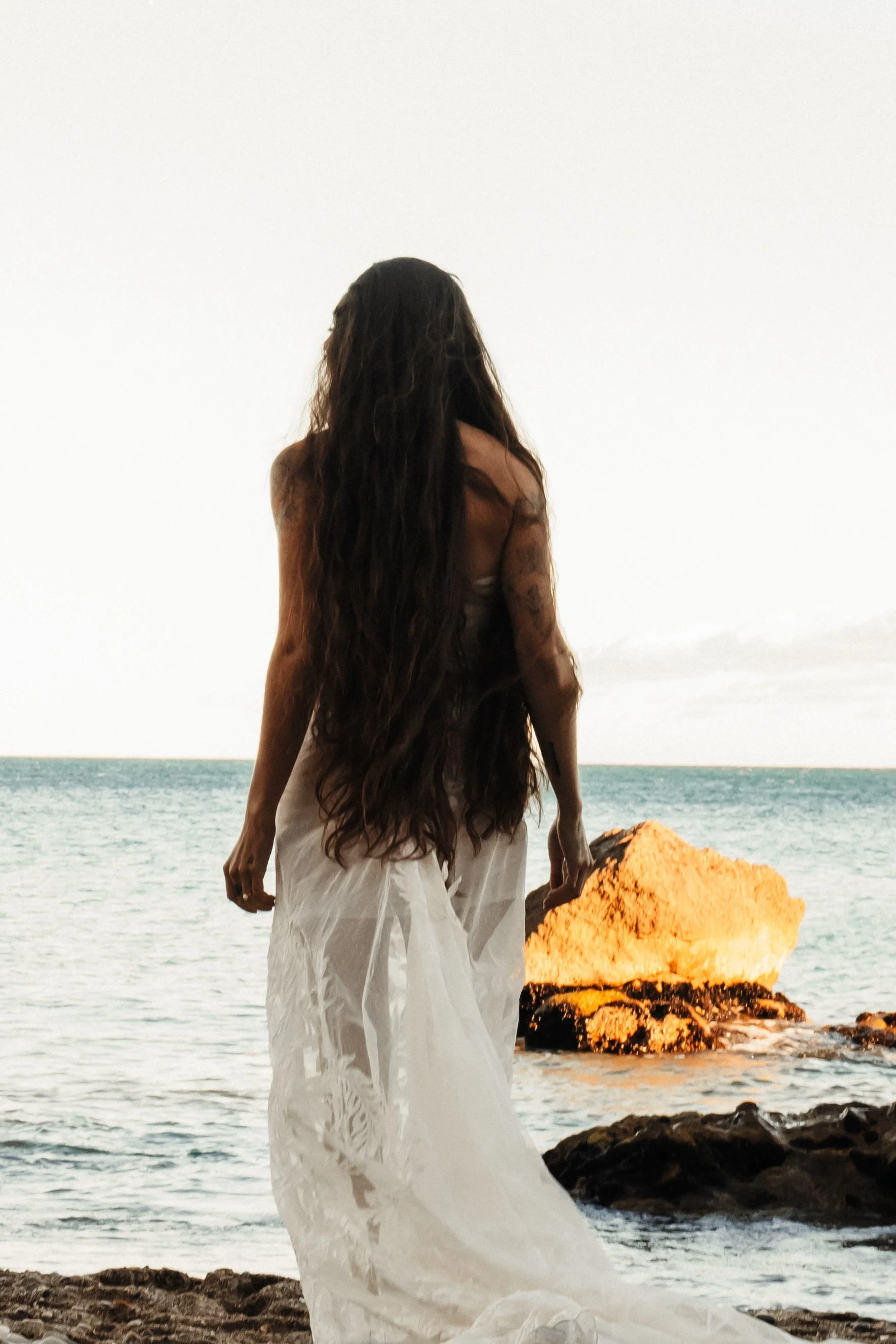 Une femme avec de longs cheveux noirs, portant une robe blanche fluide, se tient sur une plage rocheuse face à la mer au coucher du soleil.