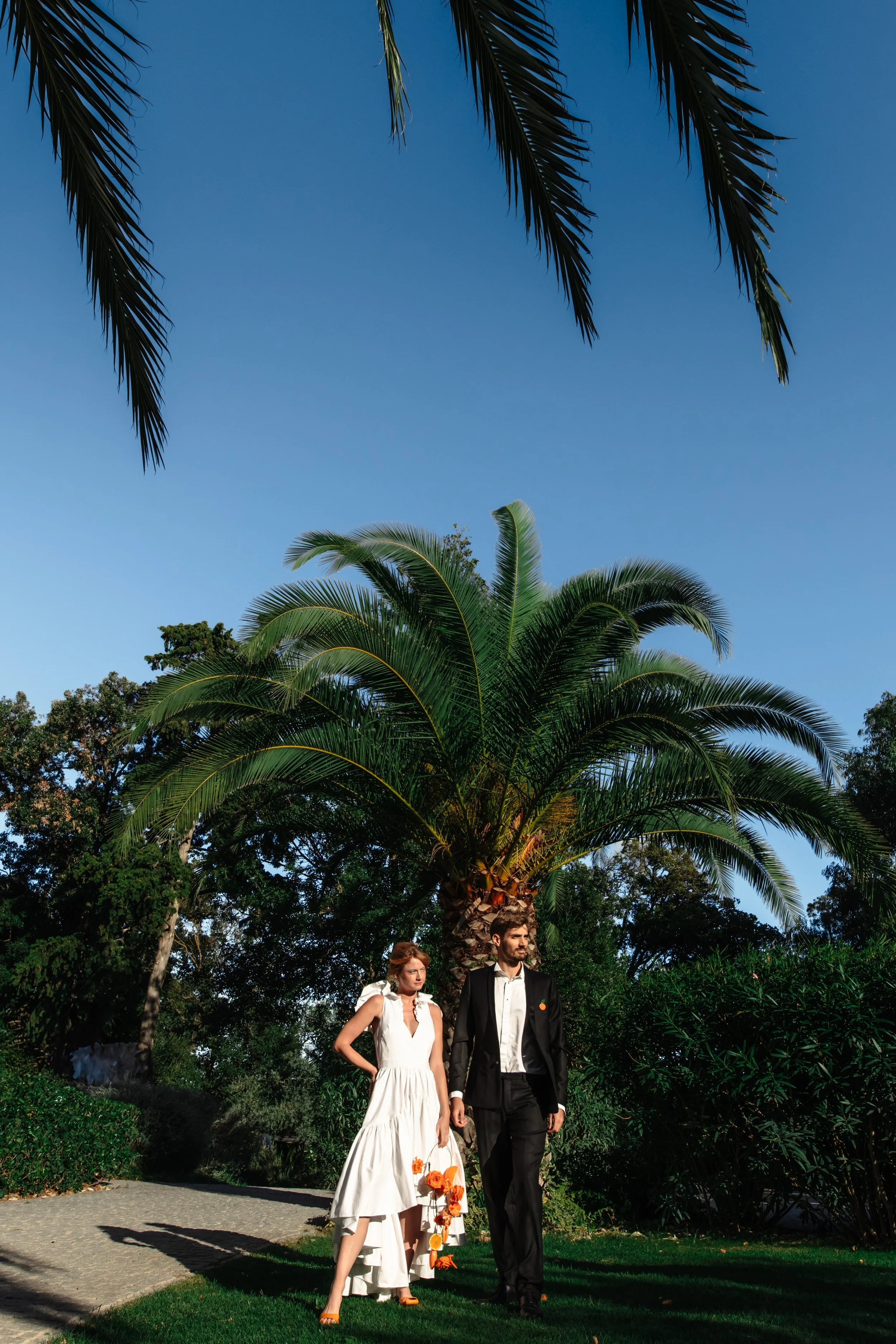 Un couple habillé élégamment marche sous un palmier dans un jardin au soleil, avec un ciel bleu clair.