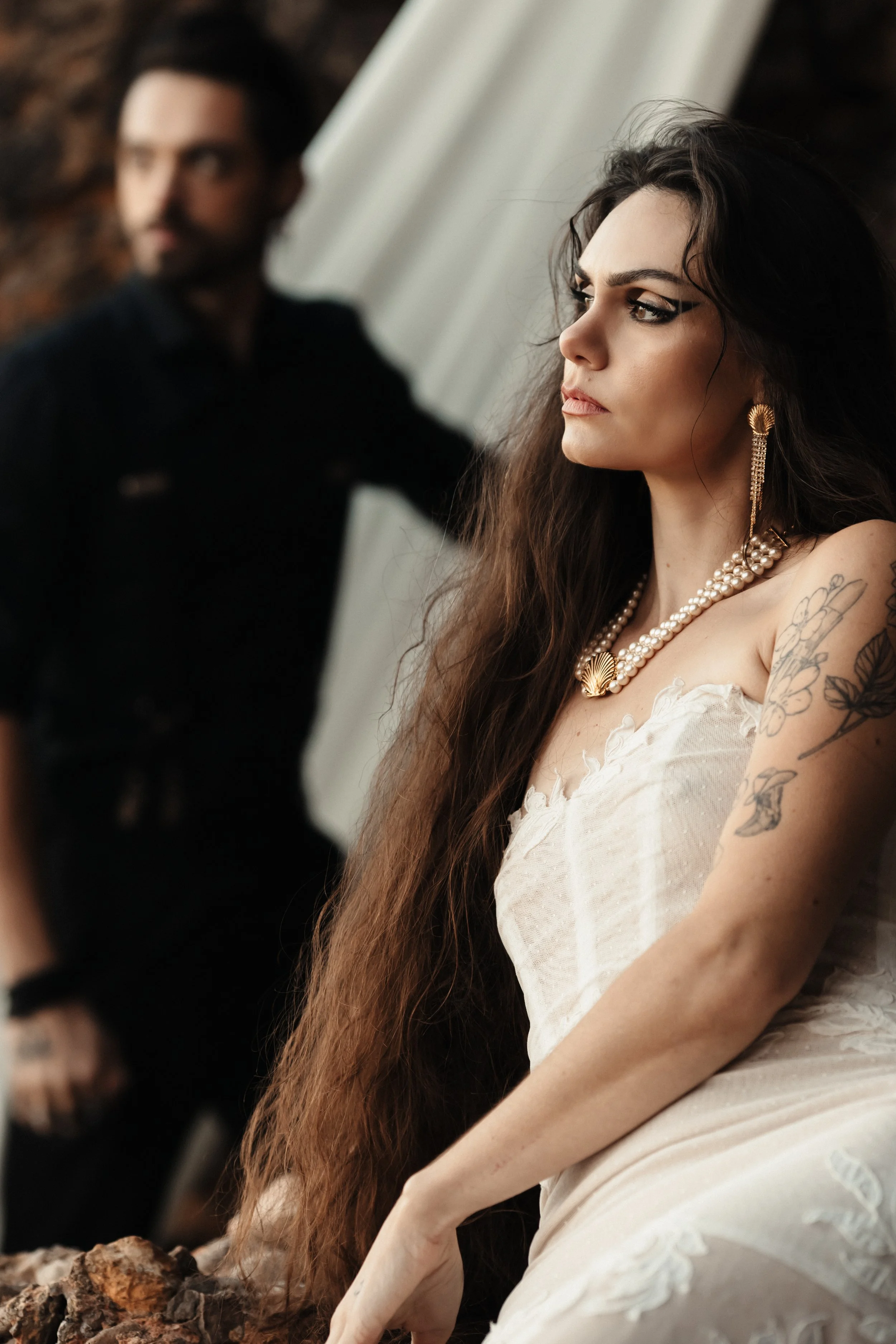 Une femme portant une robe blanche, avec de longs cheveux bruns et des tatouages sur le bras, regarde pensivement. En arrière-plan, un homme en habits noirs, flou, regarde également dans une direction différente.