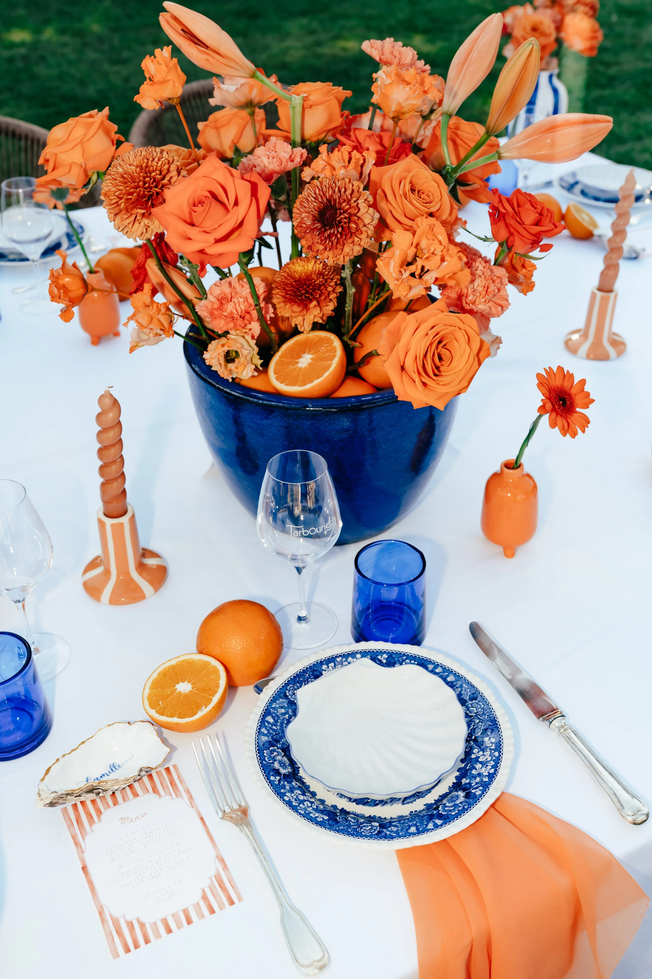 Une table de fête décorée avec un centre de fleurs orange dans un vase bleu, oranges entières et coupées, assiettes en porcelaine bleue et blanche, verre à vin, bougies, et nappe blanche avec un tissu orange.