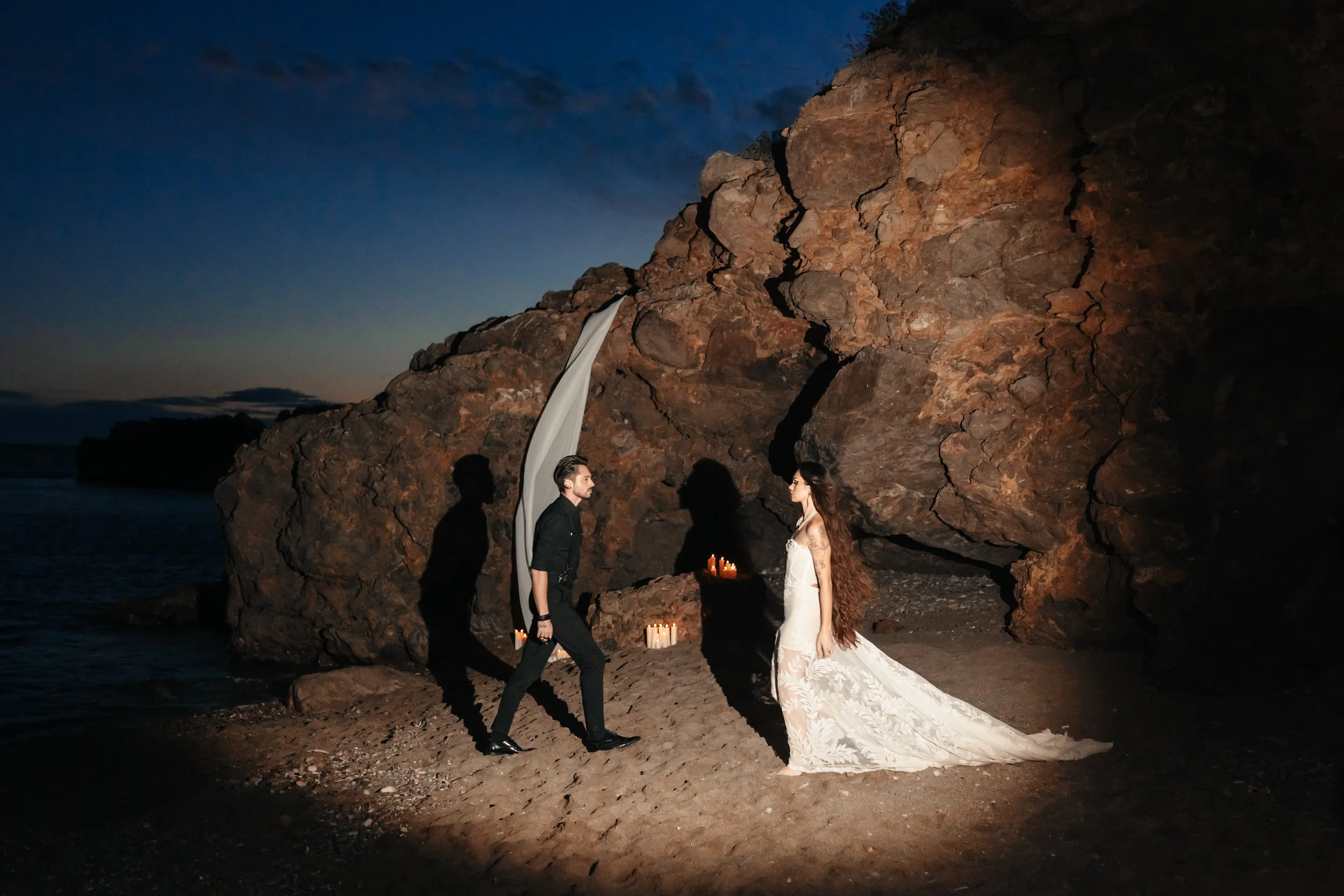 Un couple en vêtements de mariage face à face sur une plage, éclairé par des bougies, avec un rocher en arrière-plan et la mer à gauche, lors d'un crépuscule.