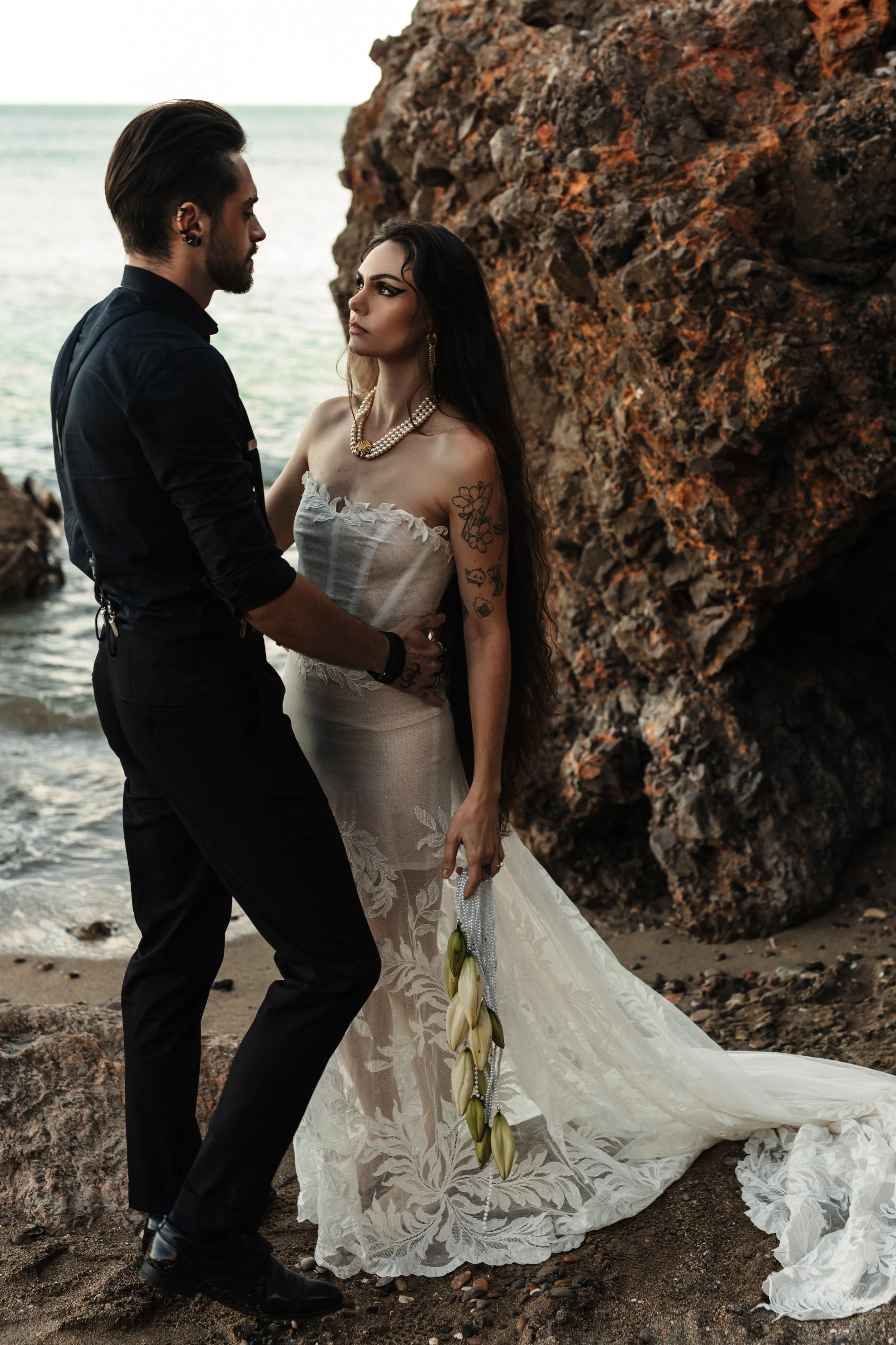 Un couple dans une scène romantique sur une plage rocheuse au coucher du soleil, la femme porte une robe de mariage blanche et tient des callaïs, tandis que l'homme, en costume noir, lève les mains pour la danser.