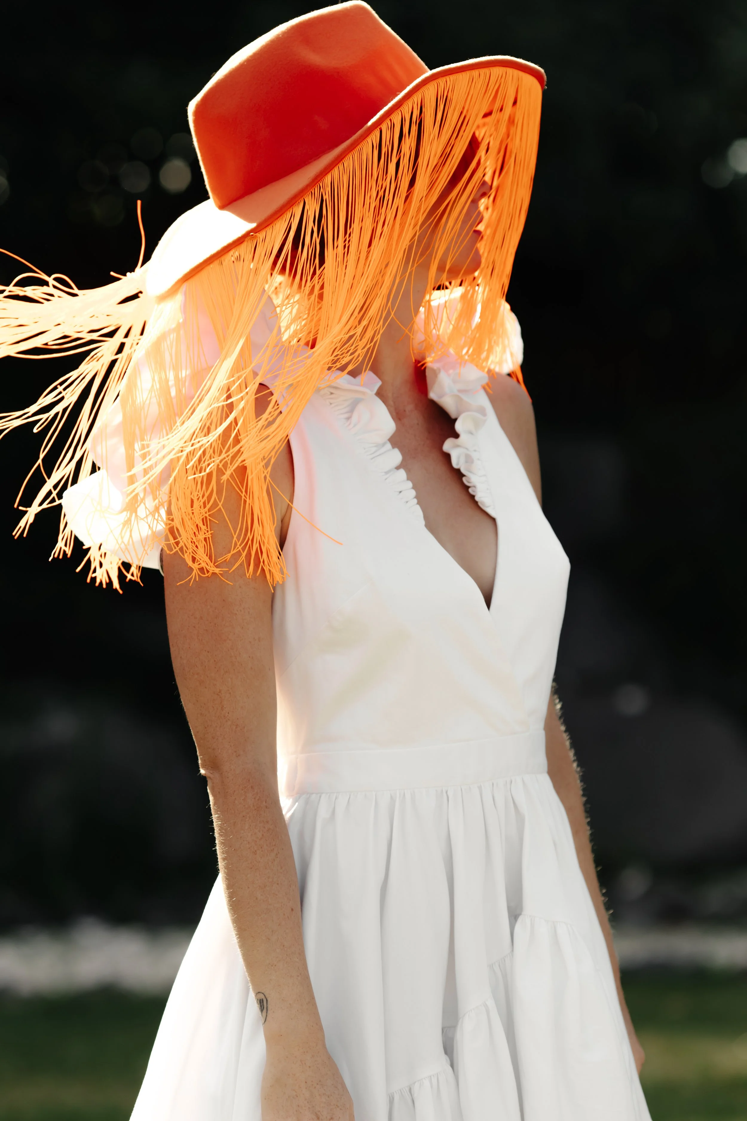 Une femme portant une robe blanche et un chapeau orange, avec un large bord, dans un décor extérieur avec un arrière-plan sombre.